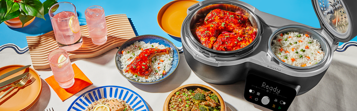 Black Dual Action multi-cooker with two open pots, one filled with saucy chicken and the other with vegetable rice, displayed on a bright, colorful table setting with plated meals, drinks, and utensils arranged around it.