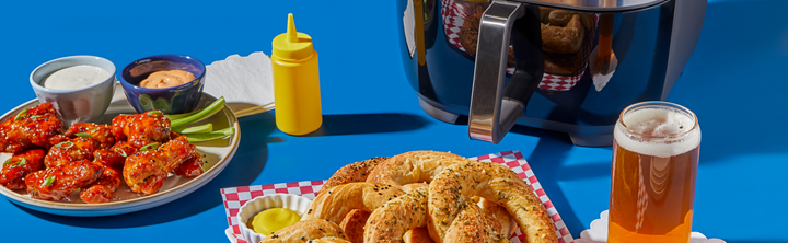 Golden, seasoned soft pretzels on a red-and-white checkered tray next to a plate of buffalo wings and a glass of beer, with an Instant Pot® air fryer and a stack of white plates in the background.
