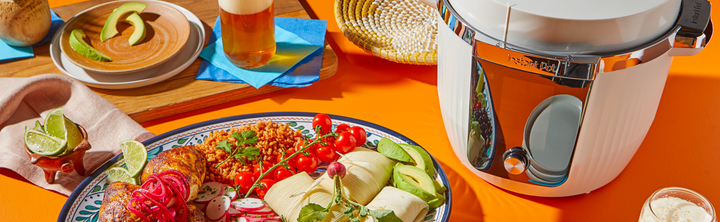 A colorful plate of tamales, rice, avocado slices, cherry tomatoes, and grilled chicken with pickled onions sits beside an Instant Pot® Pro Max on an orange table, surrounded by drinks, lime wedges, and tableware.