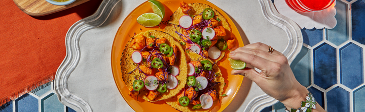 Three corn tortillas topped with seasoned chicken, sliced radishes, shredded purple cabbage, jalapeños, and green onions, served on an orange plate with lime wedges, while a hand squeezes lime juice over the tacos.