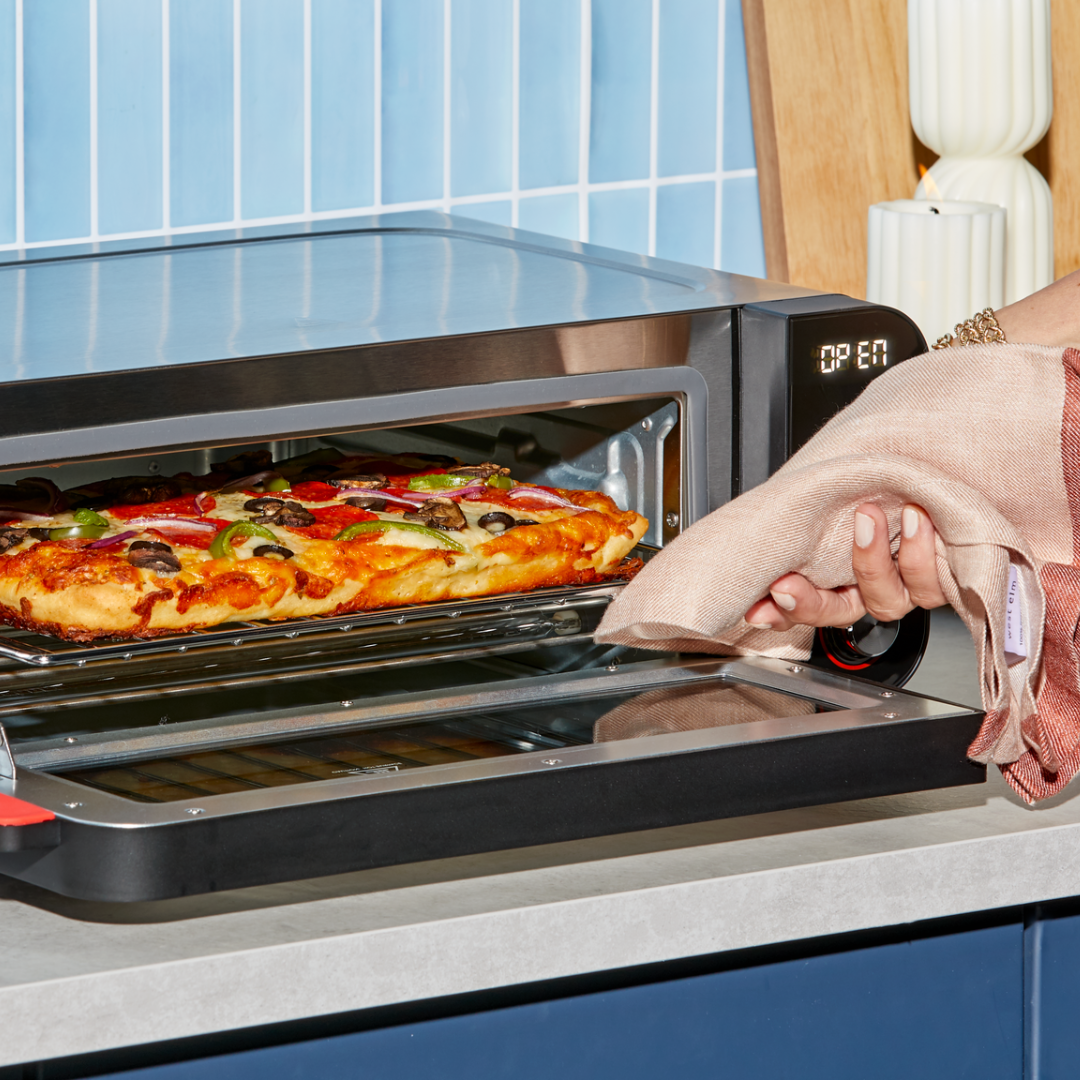 Close-up of a hand using a towel to remove a fresh, fully loaded pizza from the Instant Pot® InstantHeat™ Pizza Oven, with the display showing "OPEN" and the oven door lowered.