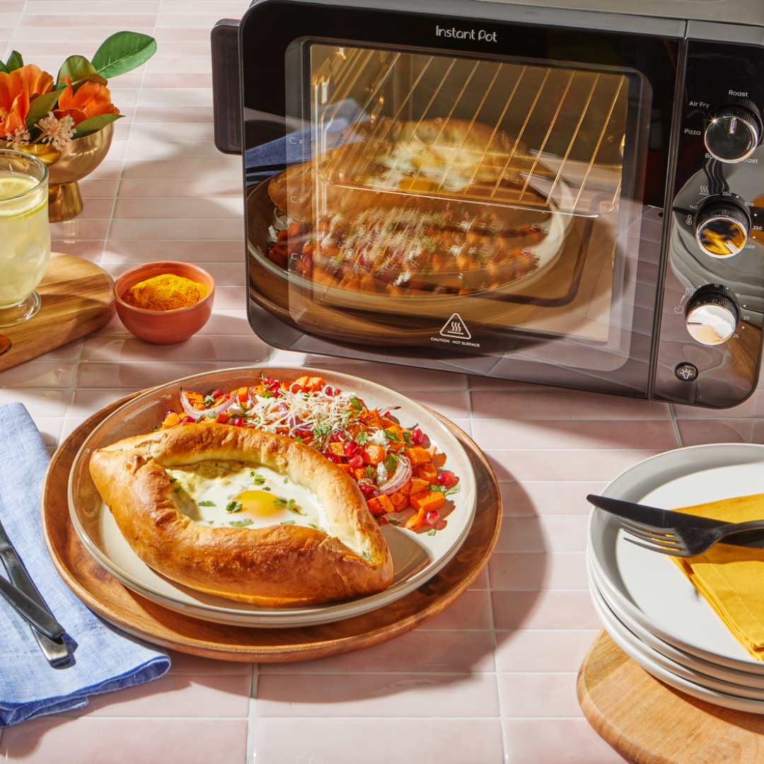 A golden khachapuri (cheese-filled bread boat with egg) served with a colorful vegetable hash sits on a plate in front of an Instant Pot® InstantHeat™ 4-Slice Air Fryer Toaster Oven. The oven is baking another khachapuri on a round tray. The scene is styled on a pink tile countertop with a drink, spices, stacked plates, and a floral arrangement nearby.