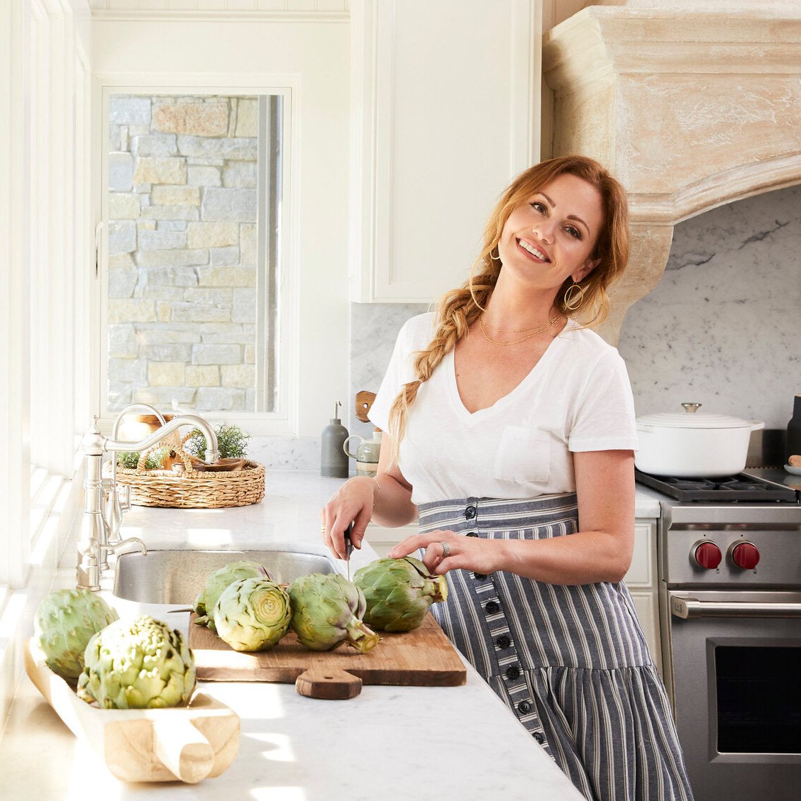 Danielle Walker, one of Instant Pot’s cooking collaborators, smiling while preparing fresh artichokes in a bright, sunlit kitchen. She wears a white t-shirt and a striped apron, with her hair styled in a loose braid.