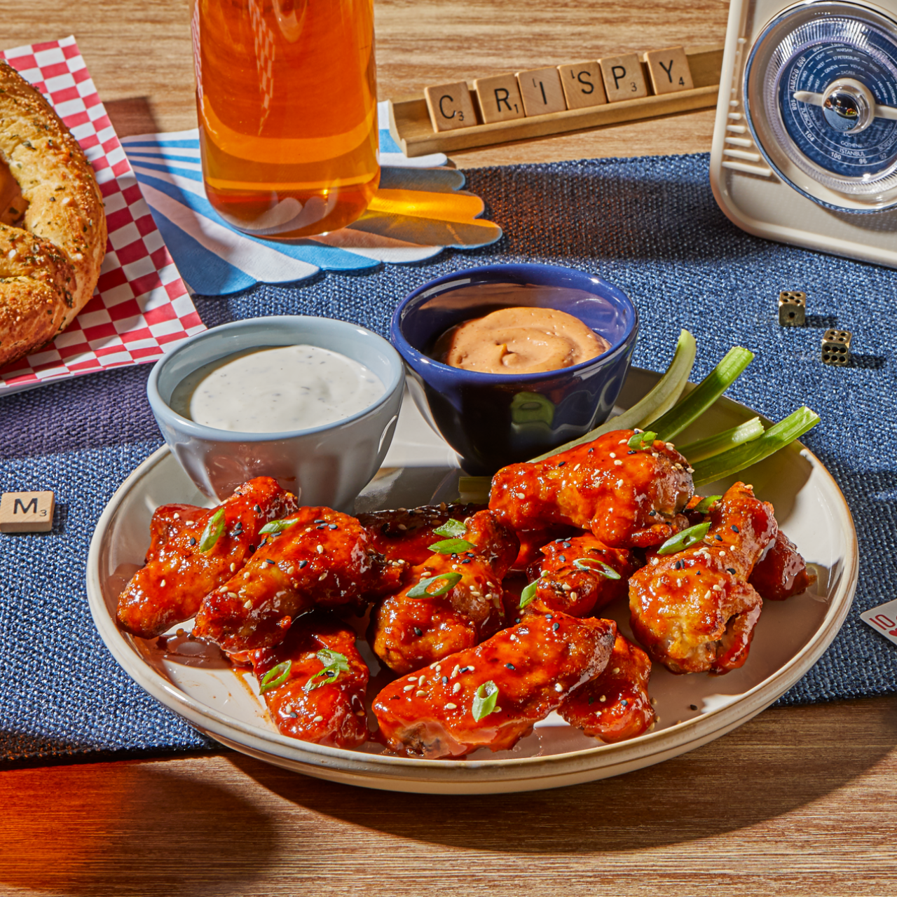 A plate of crispy, saucy chicken wings garnished with chopped scallions is served with celery sticks and two dipping sauces—one creamy white and one orange-hued—on a table set for game night. Scrabble tiles spell out "CRISPY" in the background, alongside a vintage timer, dice, a pretzel, and a glass of amber-colored drink, visually highlighting the EvenCrisp™ air frying technology's ability to deliver perfectly crunchy results.