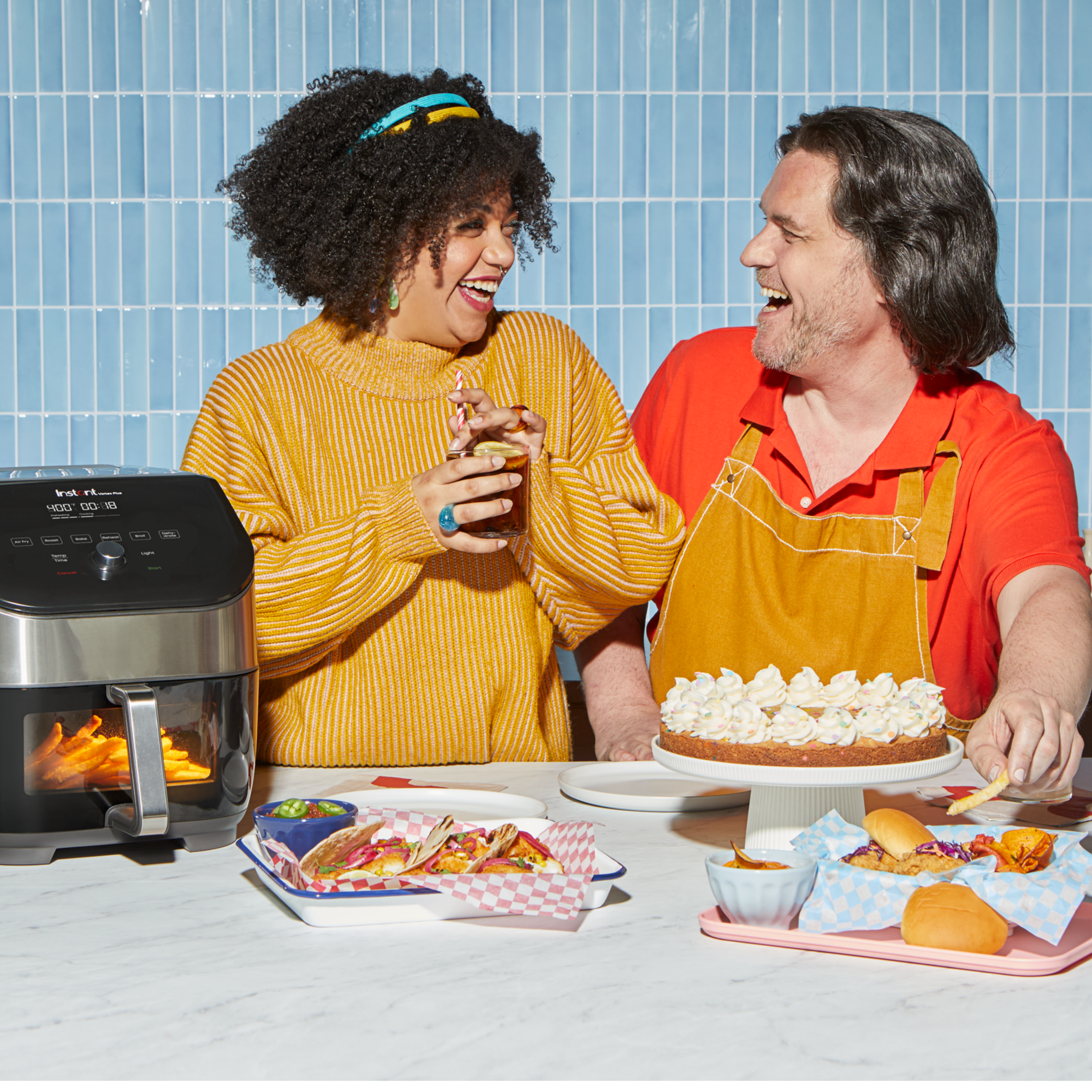 Two people laughing together in a bright kitchen setup, surrounded by plates of tacos, sliders, chips, and a frosted cake. An Instant Pot® air fryer is on the counter, mid-cook with fries inside. The woman in a yellow sweater holds a drink, while the man in a red shirt and apron reaches for a fry.