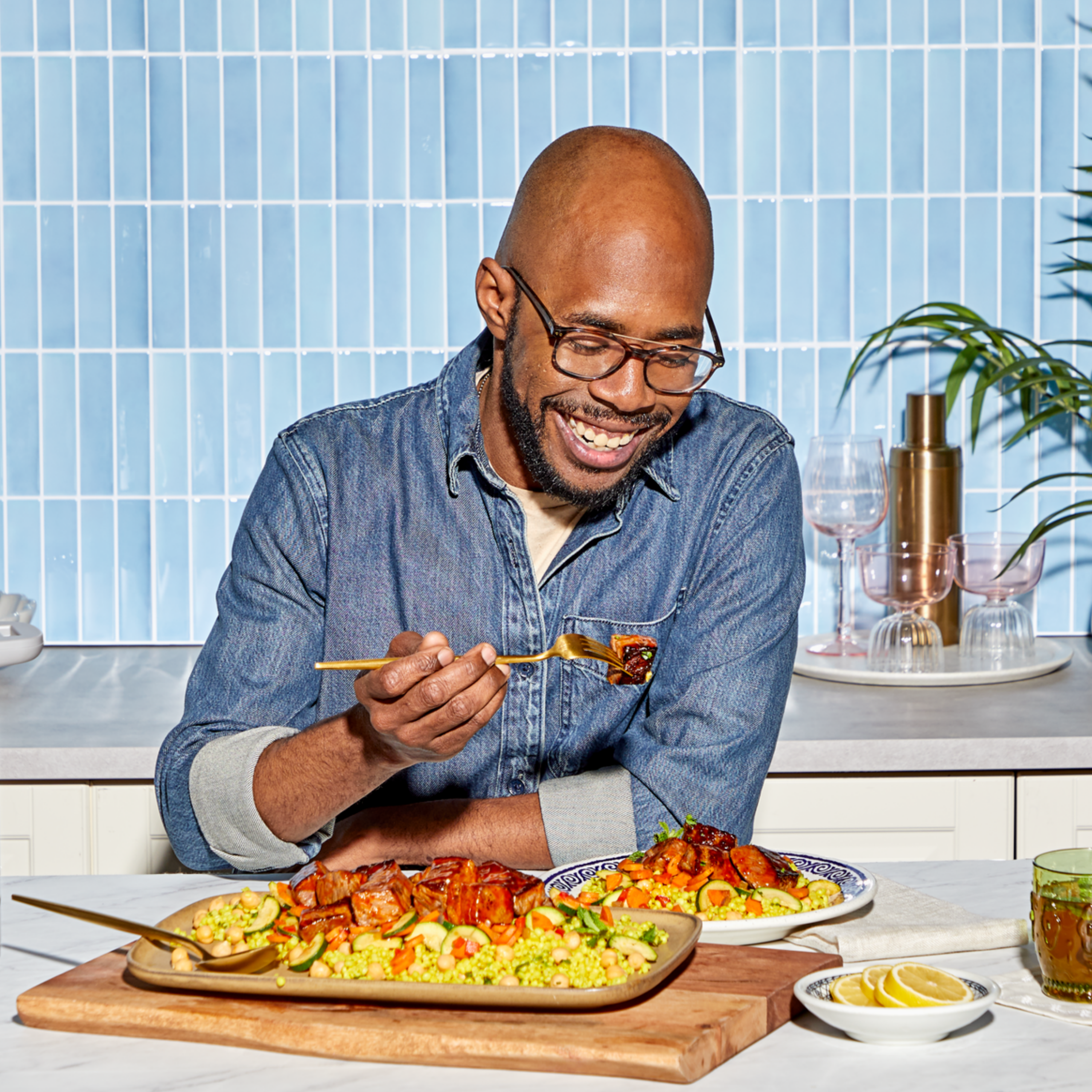 A man smiling as he enjoys a bite of food at a bright kitchen counter, dressed in a denim shirt and glasses. In front of him are colorful plates of couscous, vegetables, and glazed meat, with lemon slices and a drink nearby. A blue-tiled wall and elegant glassware add a stylish backdrop.