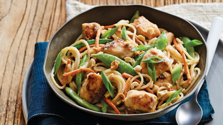 A bowl of peanut chicken and sugar snap peas with noodles, garnished with chopped peanuts, shredded carrots, and sliced green onions, served on a dark napkin with a spoon on the side.