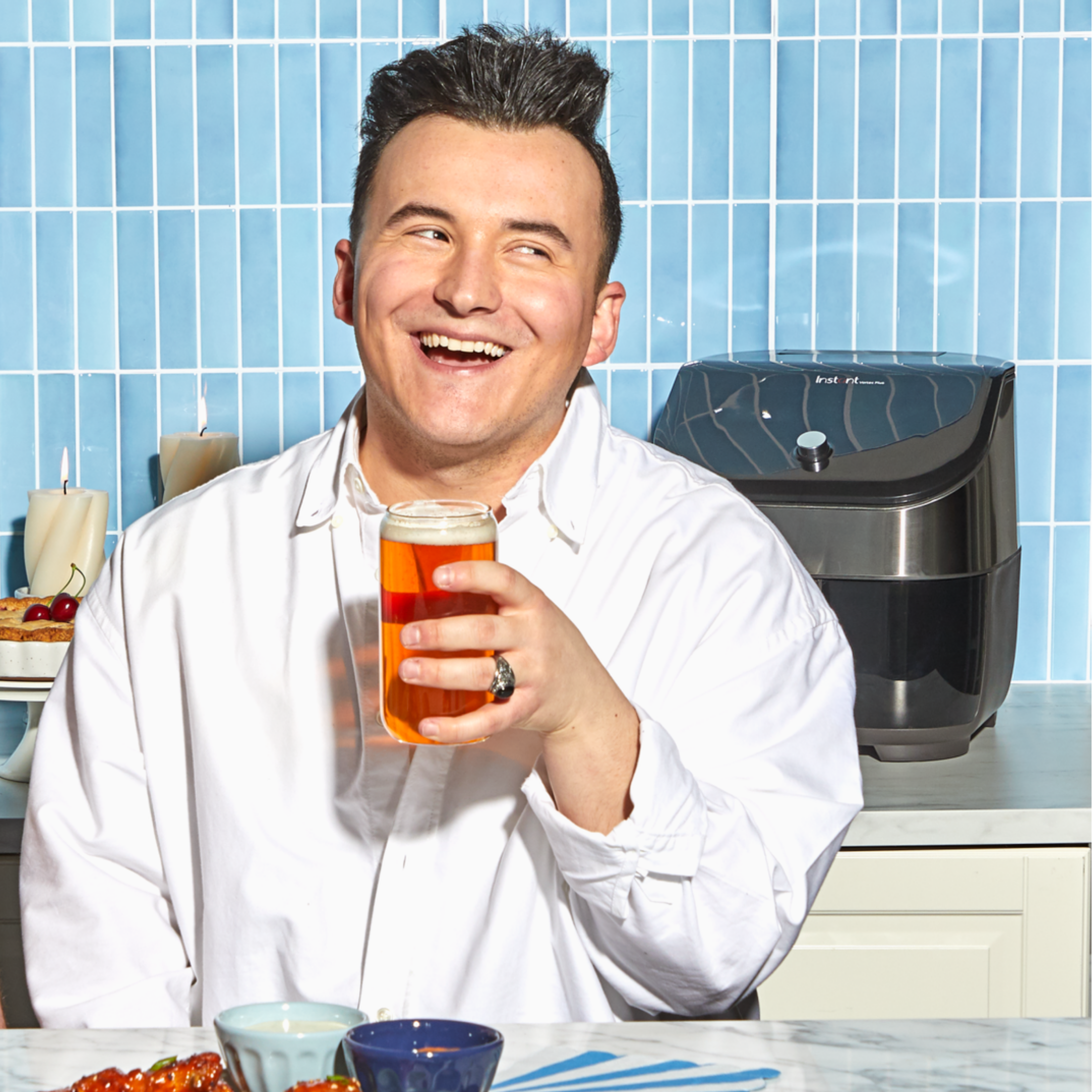 A person in a white button-up shirt smiles brightly while holding a glass of beer at a kitchen counter. In the background, an Instant Pot® air fryer sits on the counter against a bright blue tiled wall. Plates of food and dipping bowls are partially visible in the foreground.