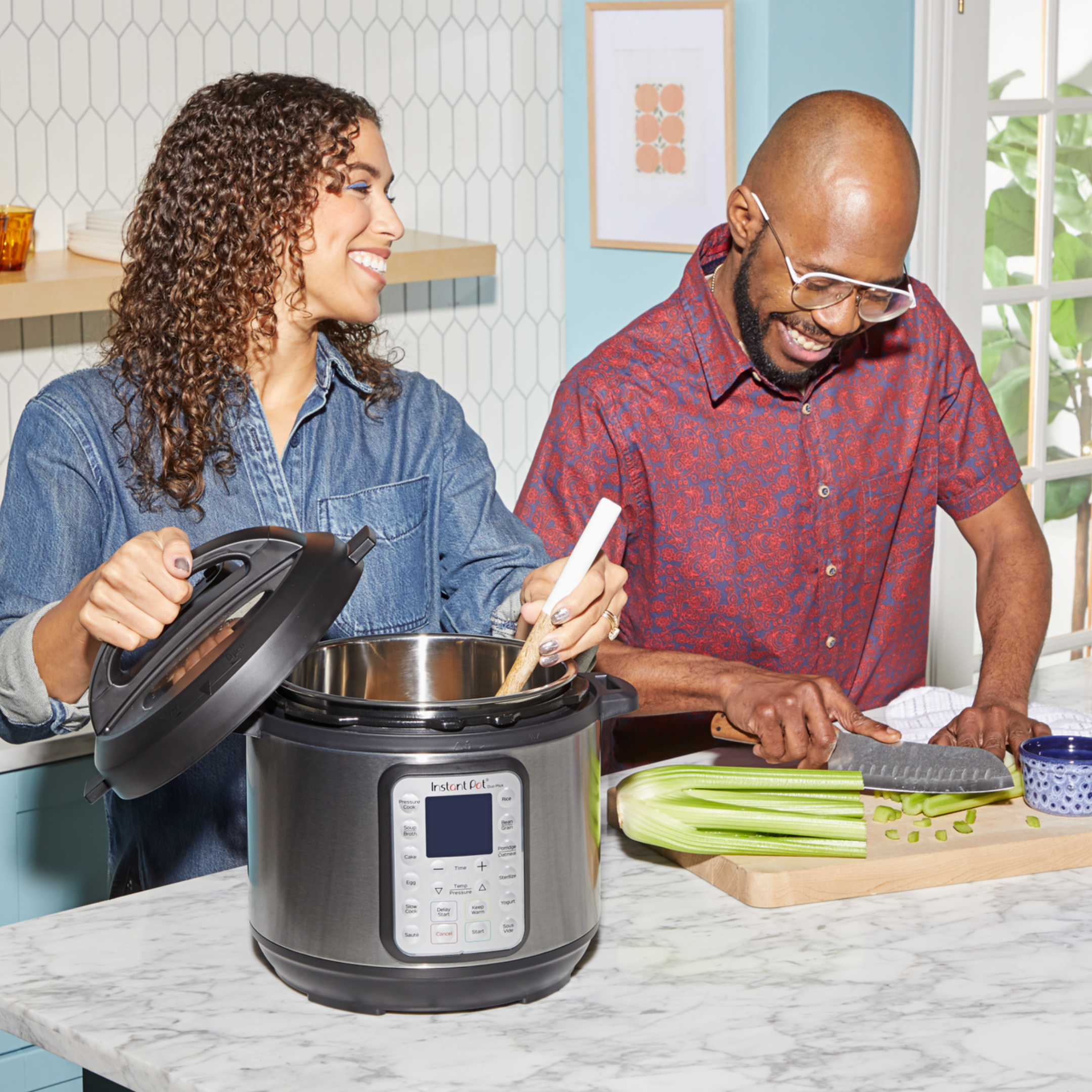A smiling couple prepares food together in a bright kitchen. One person holds the lid and stirs inside an Instant Pot® multi-cooker, while the other chops celery on a cutting board. The setting features cheerful colors, a hexagon-tile backsplash, and natural light streaming in through a nearby window.
