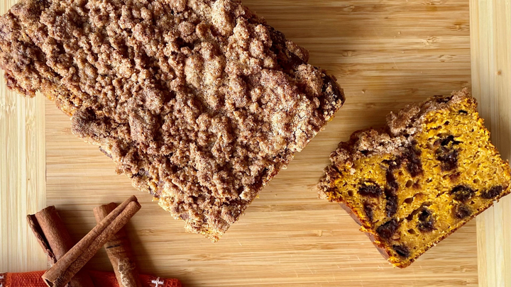 A loaf of pumpkin bread with a crumbly cinnamon streusel topping sits on a wooden cutting board beside a single sliced piece showing its golden-orange interior dotted with raisins. A few cinnamon sticks rest nearby, adding a cozy fall touch.