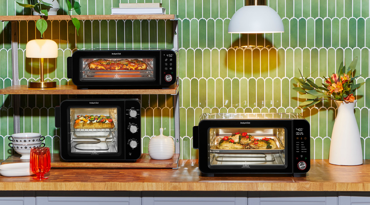 A bright, modern kitchen setup featuring three Instant Pot® countertop ovens against a glossy green tile backsplash. On the top shelf, the long, slim InstantHeat™ Pizza Oven bakes a bubbling loaf of cheesy bread. Below it, the compact, taller 4-Slice InstantHeat™ Air Fryer Toaster Oven crisps a focaccia topped with colorful vegetables. On the counter, the wide 6-Slice InstantHeat™ Air Fryer Toaster Oven roasts chicken pieces garnished with red chili slices and herbs. Warm wood tones, cozy lighting, and fres
