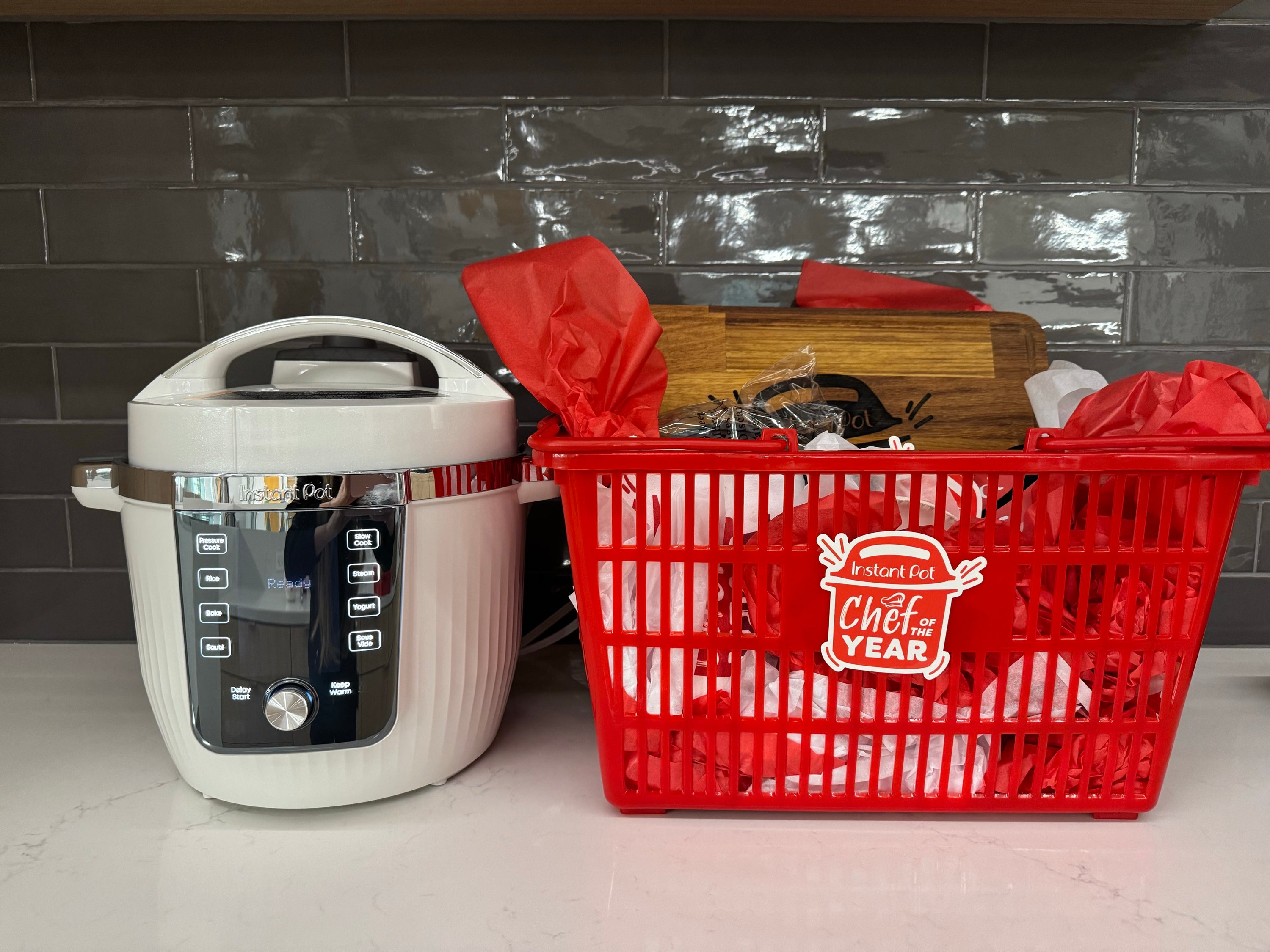 An Instant Pot Pro Max multi-cooker sits on a kitchen counter next to a red gift basket filled with tissue paper and prizes. The basket features the “Instant Pot Chef of the Year” logo and contains items including a wooden cutting board engraved with the Instant Pot logo.