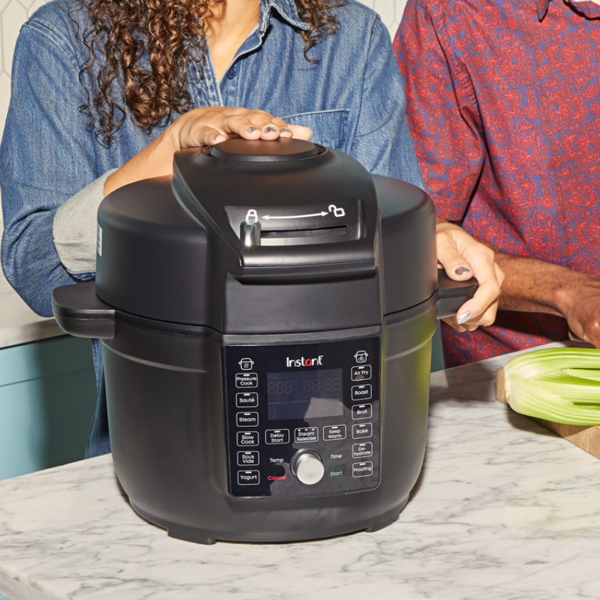 A close-up of two people using a black Instant Pot® Duo™ Crisp™ 6.5QT with Ultimate Lid Multi-Cooker and Air Fryer on a kitchen counter. One person has their hand on the pressure cooking lid, while the other holds a handle. The control panel is visible, showing various cooking functions like Pressure Cook, Air Fry, Roast, and Steam. A celery stalk sits on a cutting board nearby.