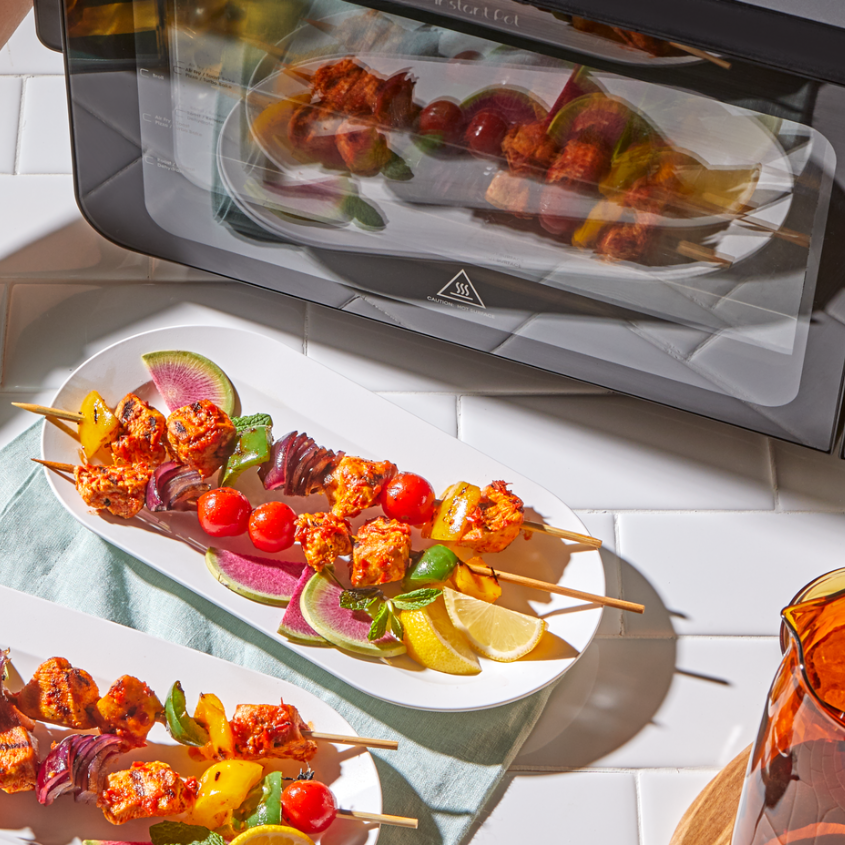 Two plates of colorful grilled skewers with chicken, cherry tomatoes, bell peppers, red onion, and watermelon radish slices are arranged on a white tiled countertop. Lemon wedges and herbs garnish the plates. In the background, an Instant Pot Toaster Oven reflects the skewers in its glass door, hinting at its recent use for cooking the meal. A pitcher of iced tea is partially visible on the side.