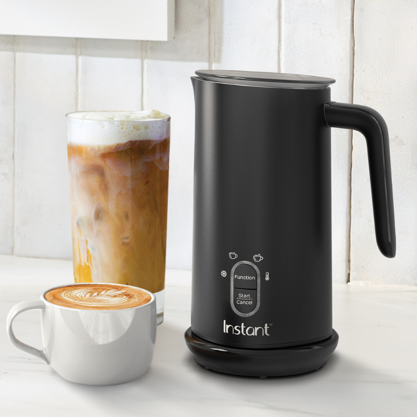 A sleek black Instant Pot Milk Frother is displayed on a white countertop next to two coffee drinks: a hot latte with latte art in a gray mug and an iced coffee with foam in a tall glass. The frother’s simple control panel features function and start/cancel buttons, emphasizing its ease of use. A white tile wall serves as the background, creating a clean, modern coffee station vibe.