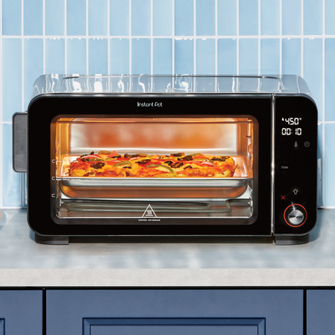 The Instant Pot® 6-Slice InstantHeat™ Air Fryer and Toaster Oven baking a rectangular pizza on the middle rack. The melted cheese and toppings are visible through the clear window, with the digital display reading 450°F. The sleek black appliance sits on a kitchen counter with a light blue tile backsplash.