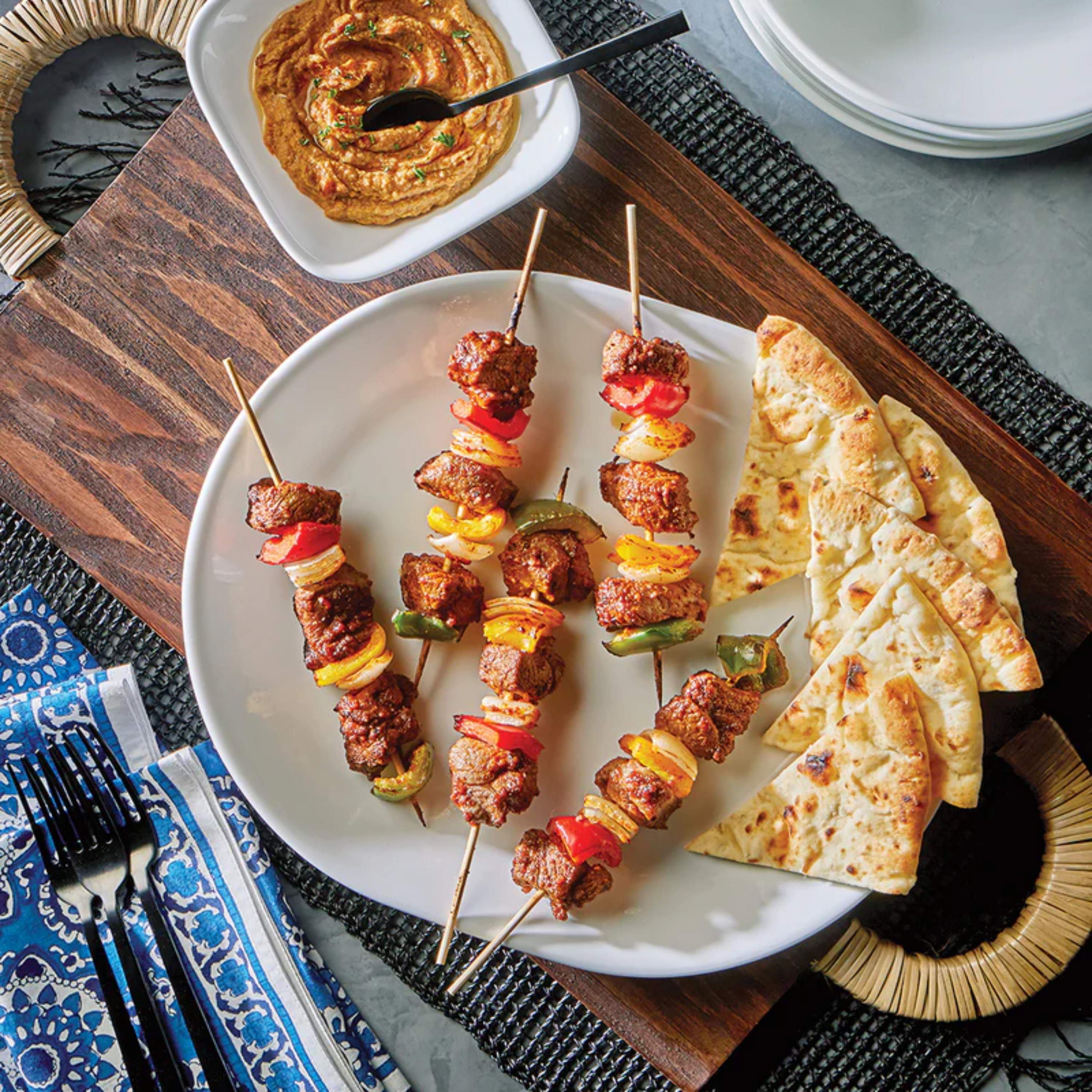 Overhead view of a Mediterranean-inspired meal featuring grilled lamb kebabs with bell peppers and onions on skewers, served with warm naan bread and a bowl of creamy hummus on a wooden tray.