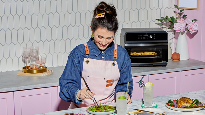 A woman in a denim shirt and light pink apron smiles while using chopsticks to eat a dish of edamame at a bright, modern kitchen counter. Behind her, an Instant Pot® toaster oven cooks fries, and the pink cabinets, floral arrangement, and stylish glassware add a cheerful, inviting vibe to the space.