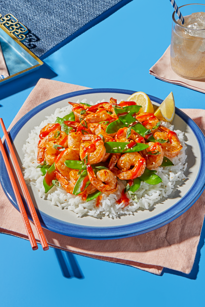 A plate of white rice topped with sautéed shrimp, snow peas, sesame seeds, and a drizzle of red sauce, garnished with lemon wedges and served with chopsticks on a blue and pink tabletop.