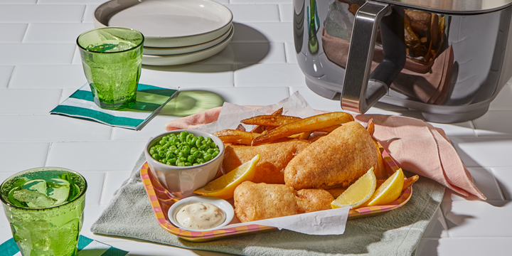 A colorful tray of air-fried fish and chips served with lemon wedges, tartar sauce, and a side of green peas. The tray is placed on a green towel next to an air fryer, green glasses of ice water, and a stack of plates on a white tiled surface.