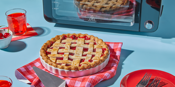 A freshly baked cherry pie with a golden lattice crust sits on a red checkered towel in front of an Instant Pot toaster oven. The scene includes a red drink with ice, a bowl of fresh cherries, a red plate with forks, and a pie server, all set on a light blue surface for a vibrant, summery look.
