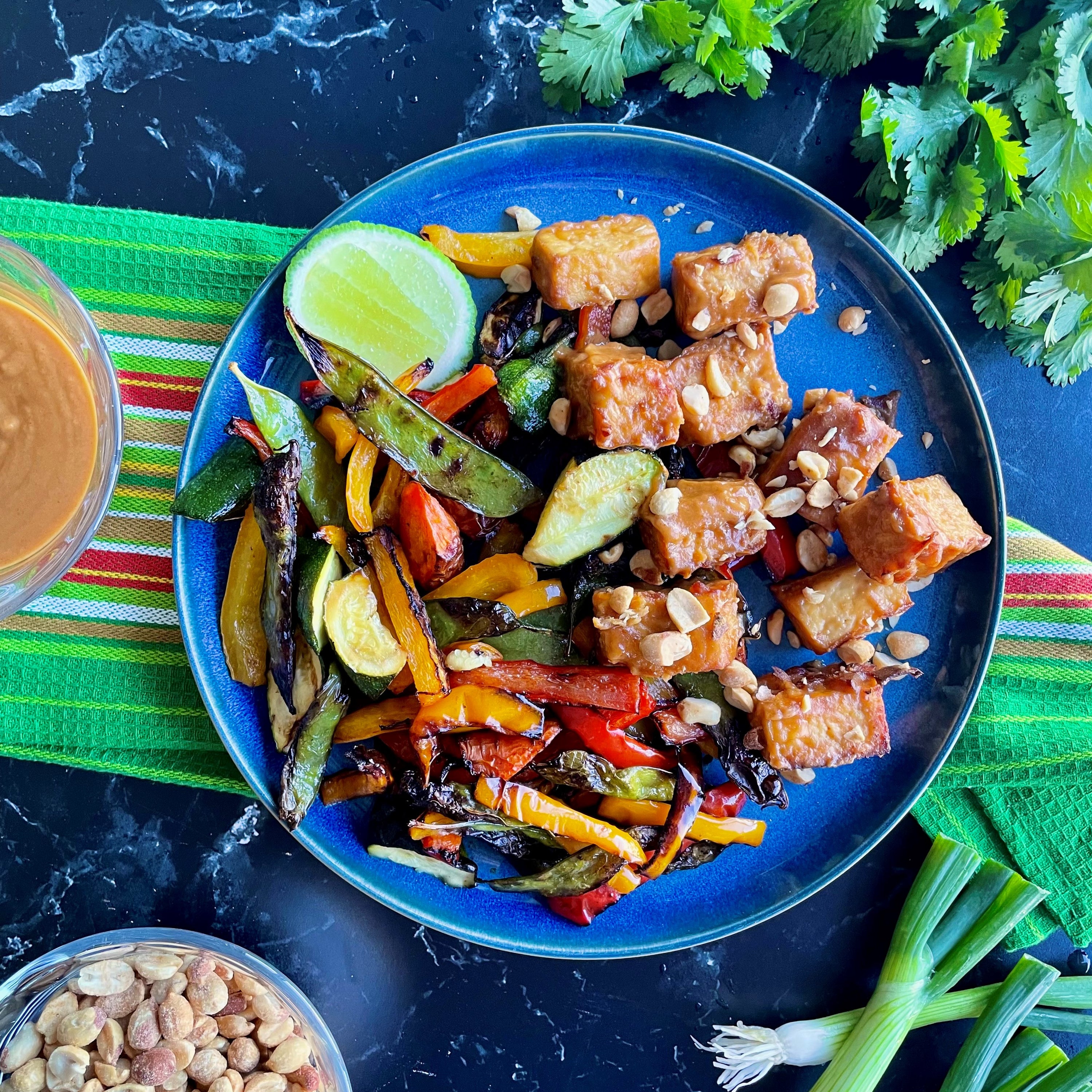 Colorful Thai Peanut Tofu dish on a blue plate, featuring golden-brown tofu cubes topped with chopped peanuts, served alongside stir-fried vegetables like bell peppers, zucchini, and snow peas. A lime wedge is placed on the side. Recipe by Instant Pot Culinary Team.