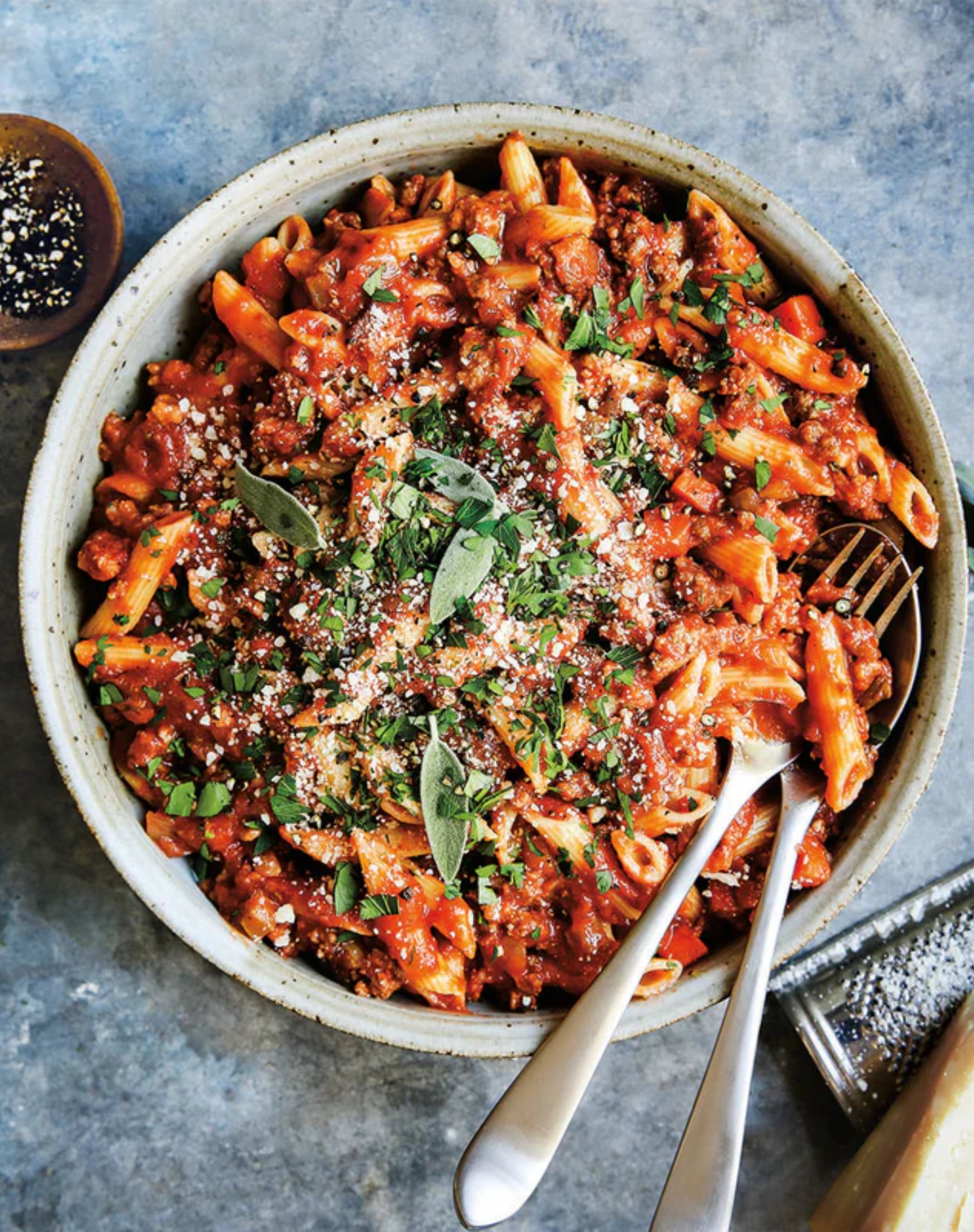 A rustic bowl filled with penne pasta coated in a rich Bolognese sauce, topped with chopped parsley, grated Parmesan, and fresh sage leaves, with two forks nestled into the dish on a textured blue-gray surface.