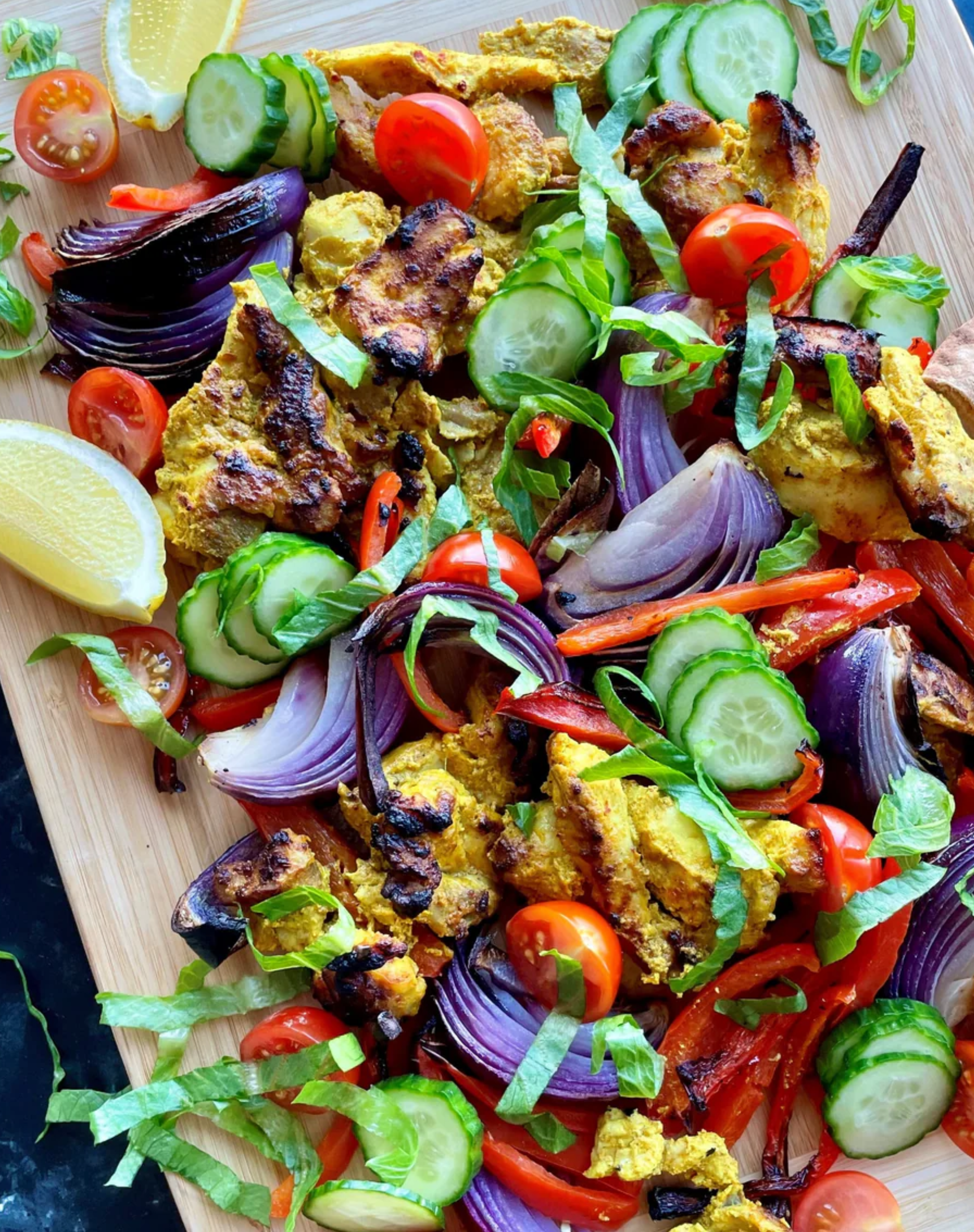 A vibrant spread of grilled chicken shawarma on a wooden board, surrounded by charred red onions, sliced cucumbers, cherry tomatoes, red bell peppers, shredded lettuce, and lemon wedges for garnish.