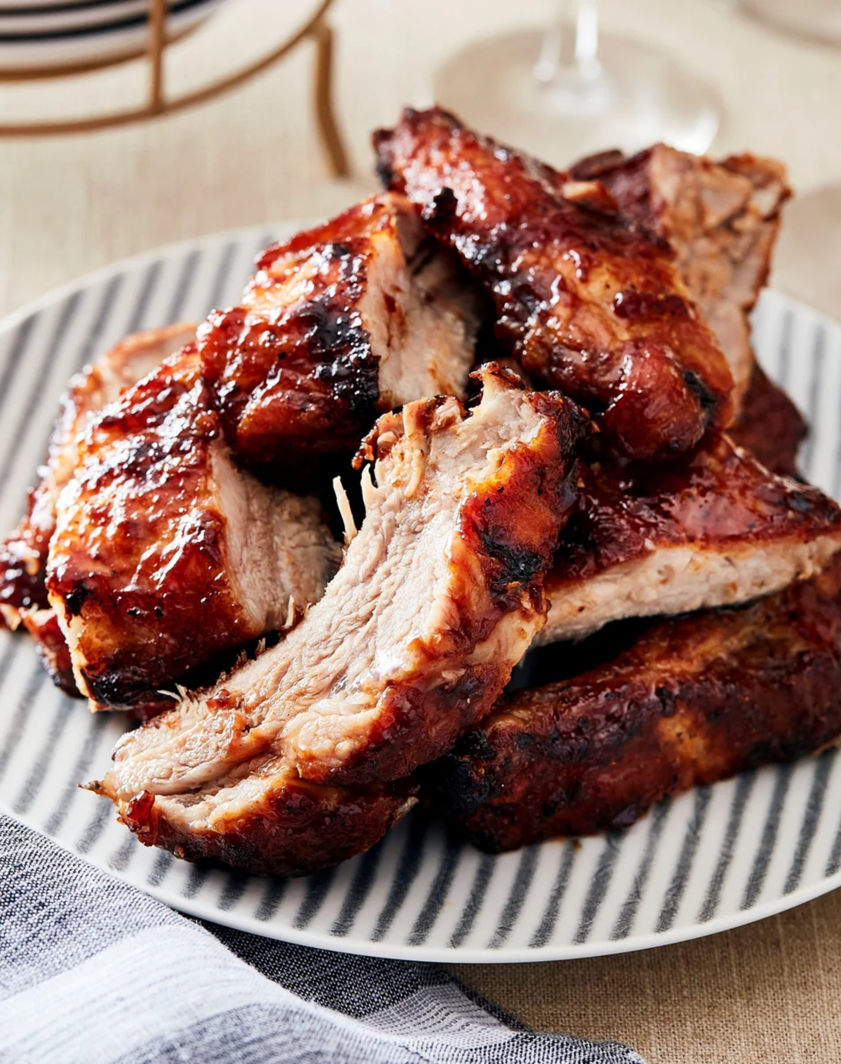 A plate stacked high with juicy, glazed baby back ribs, showing off their caramelized crust and tender, fall-apart interior, served on a striped dish with a cloth napkin nearby.