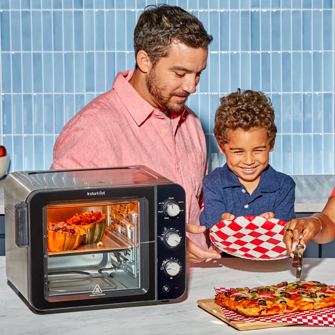 A smiling man and young boy stand beside the Instant Pot® InstantHeat™ 4-Slice Air Fryer Toaster Oven, which is cooking stuffed squash. The boy holds a red-and-white checkered plate while a hand slices a veggie-topped pizza on a nearby cutting board. The setting is a bright, modern kitchen with a blue tile backsplash.
