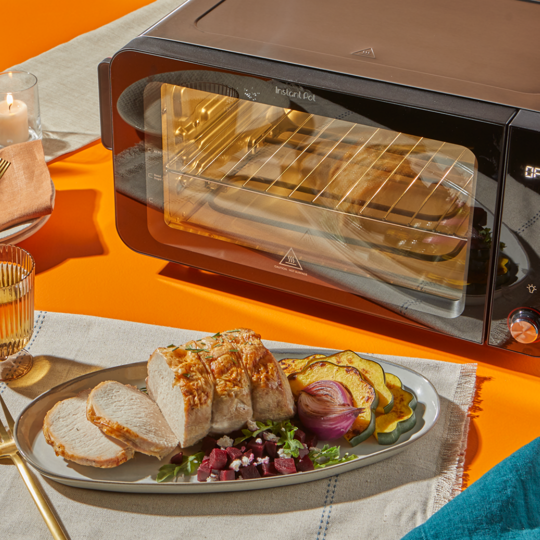 The Instant Pot® InstantHeat™ 6-Slice Toaster Oven sits on a bright orange tabletop with its oven light on and food baking inside. In front of it, a plated meal of sliced roasted chicken breast, grilled acorn squash, red onion, and beet salad is served on a linen runner alongside a gold fork and a glass of water.