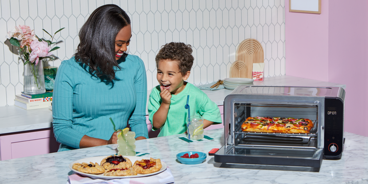 A smiling woman in a teal top sits next to a laughing child at a kitchen counter. Both have drinks with curly straws in front of them, along with a plate of mini fruit galettes and a bowl of raspberries. To their right, the Instant Pot® 6-Slice InstantHeat™ Air Fryer and Toaster Oven is open, displaying a baked rectangular pizza inside. The kitchen features a white tile backsplash, pink cabinets, and cheerful decorative accents, creating a warm and playful atmosphere.