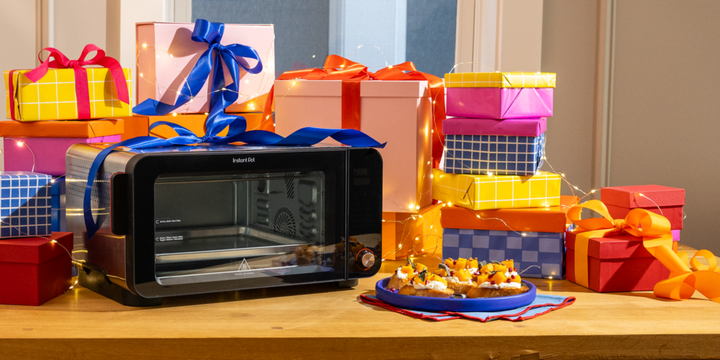 Instant Pot® InstantHeat™ 6-Slice Air Fryer Toaster Oven displayed on a wooden counter, surrounded by colorful wrapped gift boxes and string lights. A plate of festive appetizers sits beside the oven, creating a bright holiday scene.