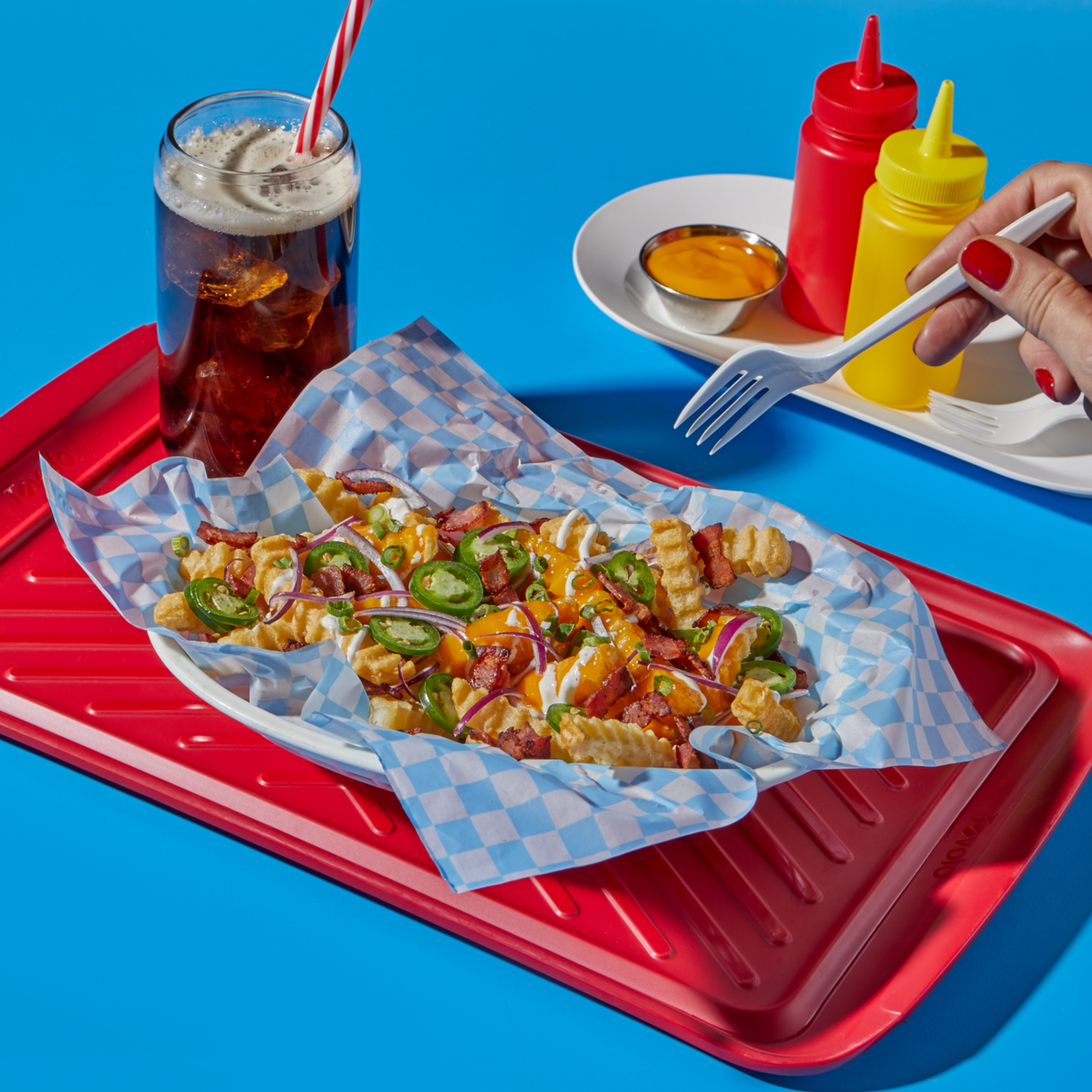 A tray of loaded crinkle-cut fries topped with melted cheese, sliced jalapeños, bacon, red onions, and a drizzle of sauce, served on blue checkered paper. A hand with red nail polish holds a white fork near the tray. In the background, there’s a glass of soda with a red-striped straw and a white condiment tray with ketchup, mustard bottles, and a cup of cheese sauce, all set on a bright blue background.