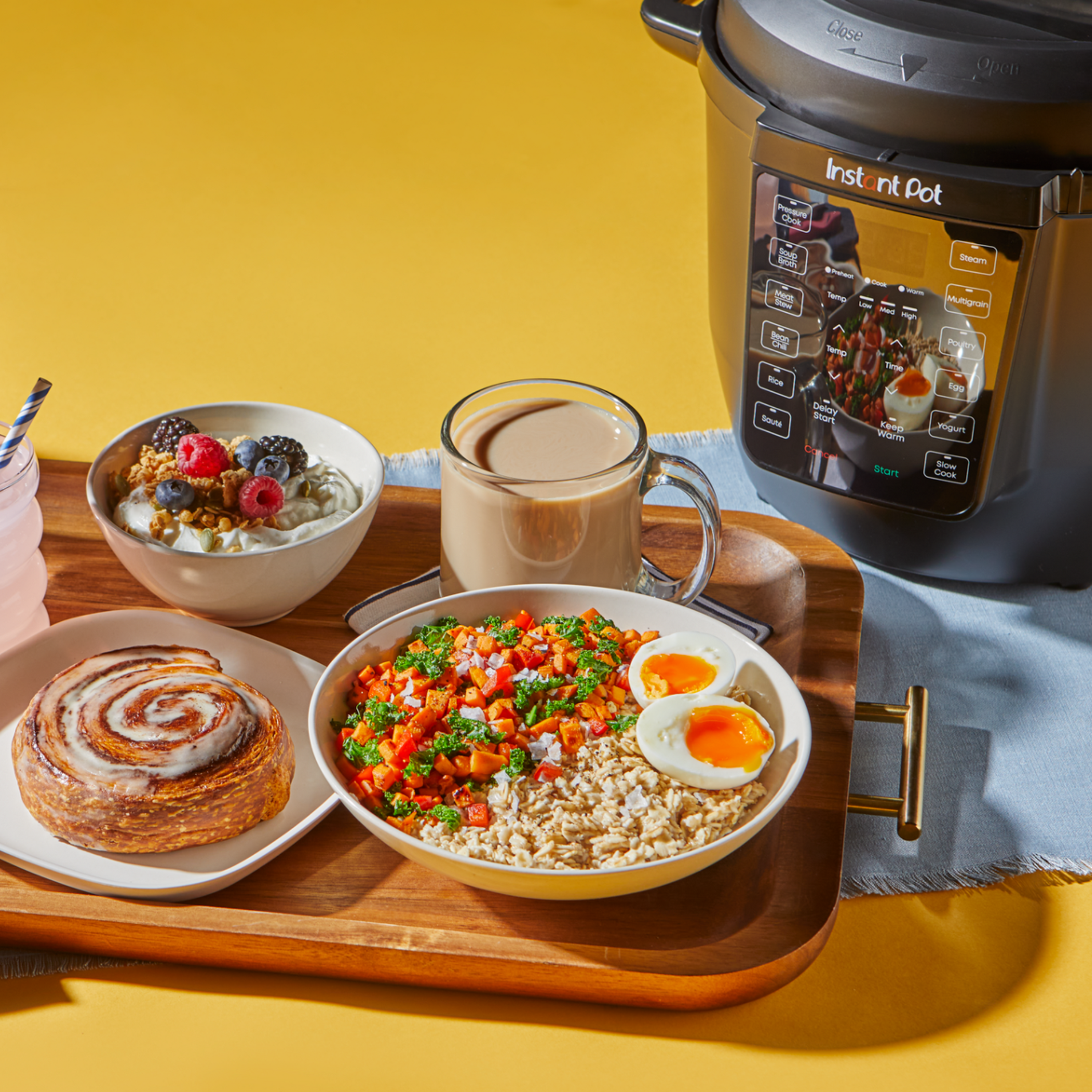 A hearty breakfast spread is served on a wooden tray in front of an Instant Pot multi-cooker. The tray includes a bowl of brown rice topped with sautéed vegetables and two soft-boiled eggs, a glass mug of coffee, a cinnamon roll on a plate, and a bowl of yogurt topped with granola, raspberries, blackberries, and blueberries. A pink drink with a striped straw is placed next to the tray. The Instant Pot is shown in the background on a light blue cloth against a yellow backdrop.