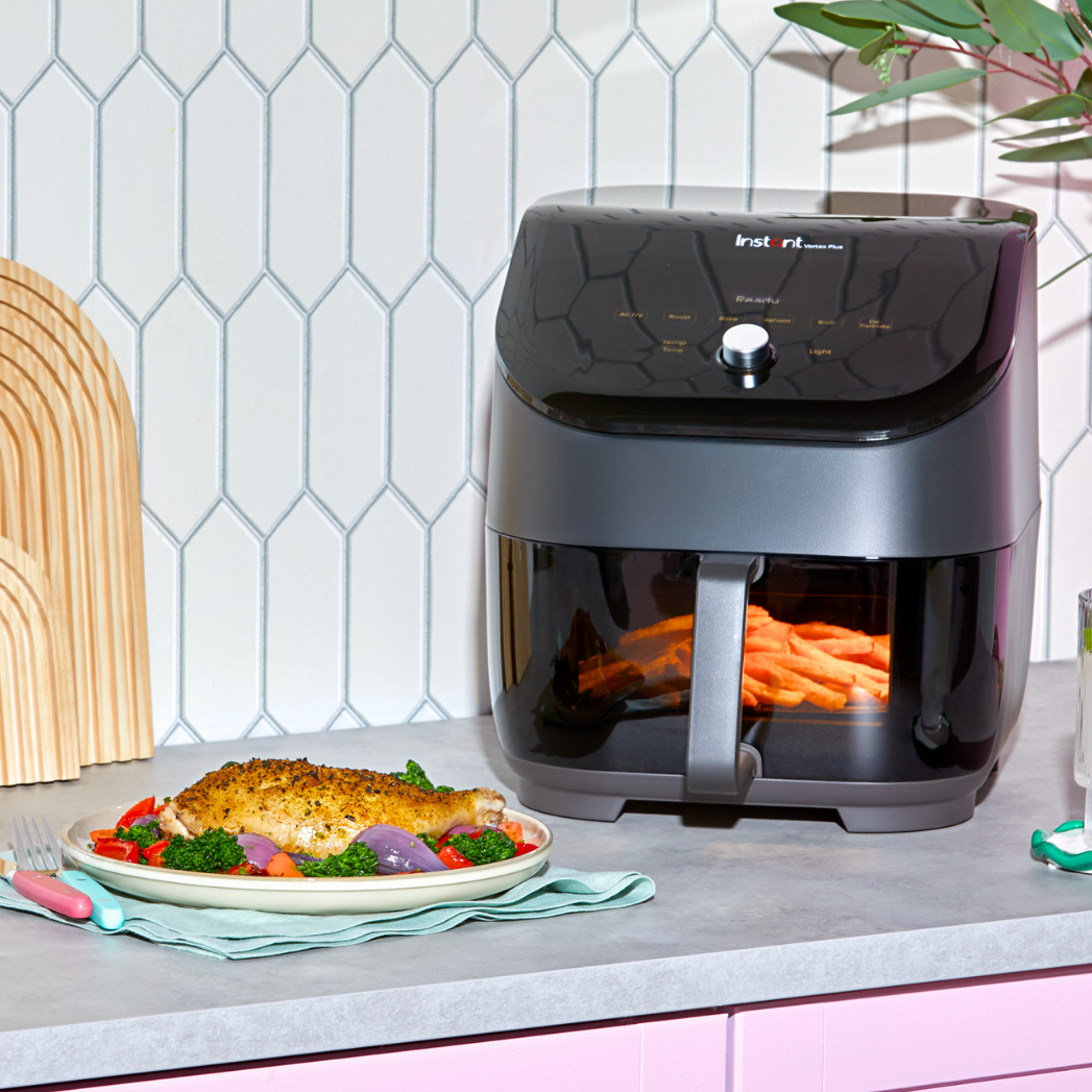 An Instant Pot® Vortex® air fryer with a visible basket of fries sits on a gray kitchen counter in front of a white geometric tile backsplash. Nearby, a plate with a breaded fish fillet, broccoli, and roasted vegetables is set on a pastel green napkin with utensils.