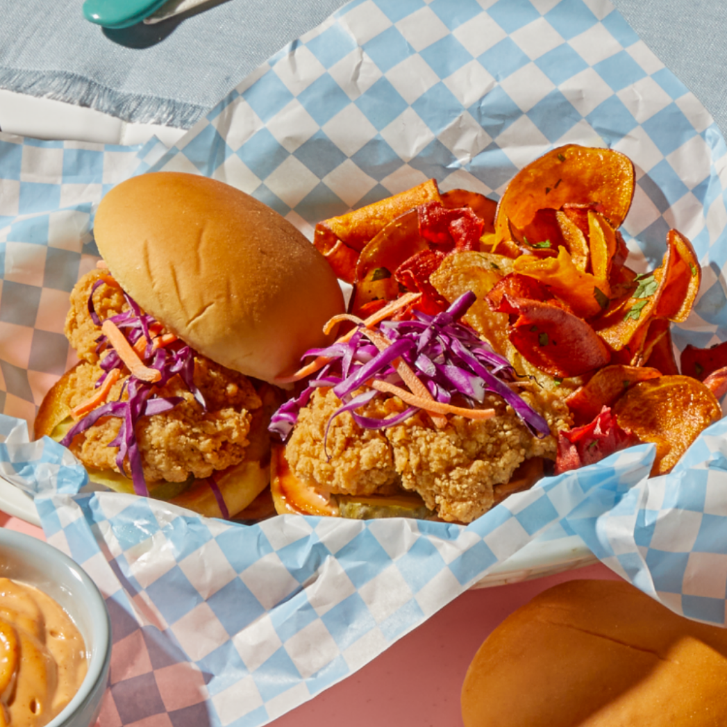  A checkered blue-and-white paper-lined tray holds two fried chicken sandwiches topped with shredded purple cabbage, served with a side of colorful vegetable chips. A bowl of dipping sauce and an extra bun are partially visible nearby.