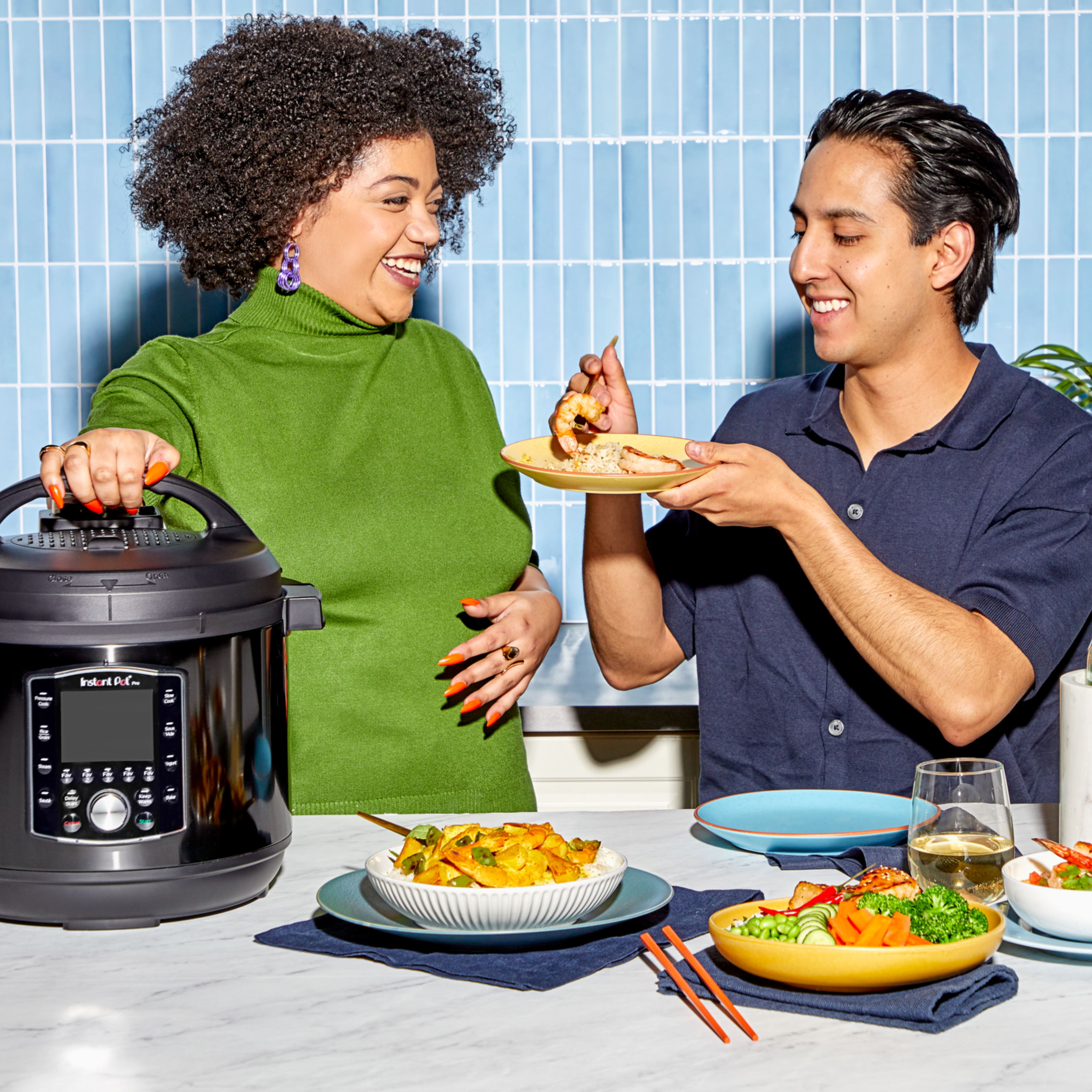 Two people share a joyful moment while cooking and eating together in a brightly lit kitchen. The woman in a green sweater rests her hand on an Instant Pot multi-cooker, smiling at the man in a navy shirt who is holding a plate and offering her a bite of food. On the table in front of them are plates of colorful dishes, including rice with vegetables, sliced cucumbers and carrots, and a glass of white wine. The backdrop features a blue tiled wall and a modern kitchen setting.