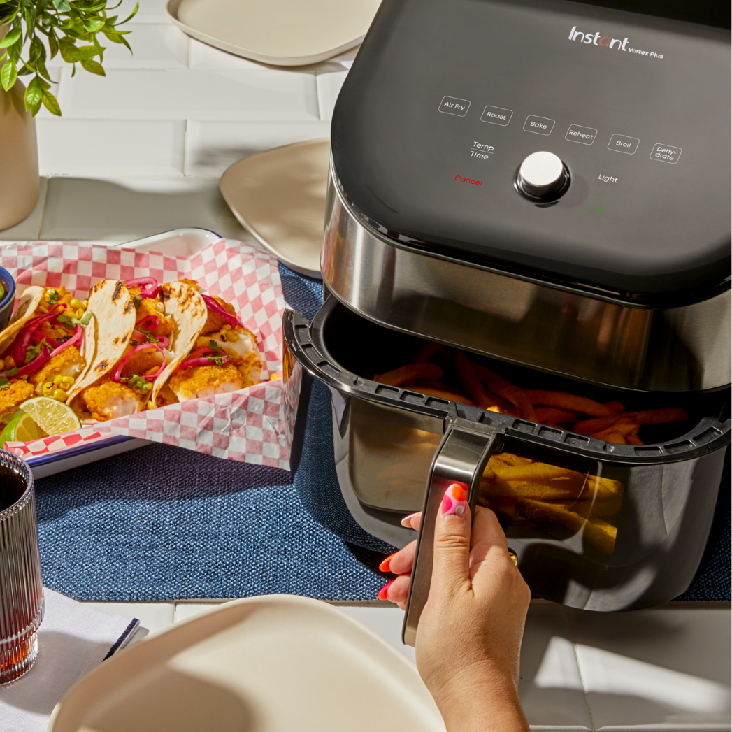 A person opens the basket of an Instant Pot® Vortex® air fryer filled with fries, next to a plate of tacos in a red-and-white checkered tray on a blue placemat. Plates, a drink, and a potted plant are visible on a white tiled table.