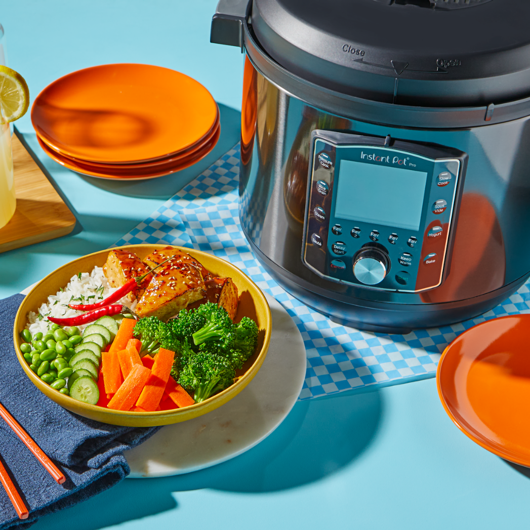 A colorful bowl filled with rice, soy glazed chicken, broccoli, carrots, cucumber, peas, and a red chili sits in front of a black Instant Pot® multi-cooker on a bright blue surface. Orange plates, a blue napkin with chopsticks, and a glass of lemonade with lemon slices complete the cheerful setup.