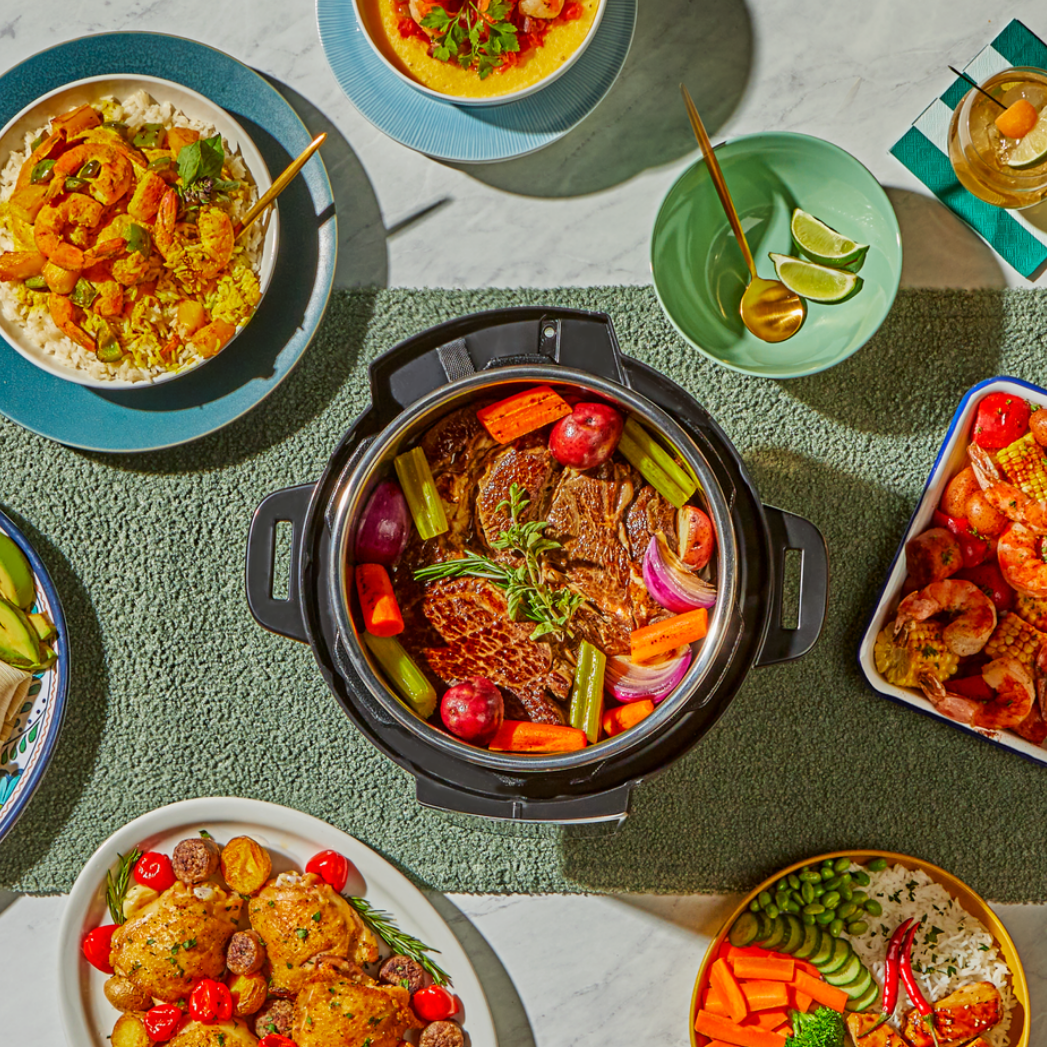An overhead view of a feast featuring an Instant Pot® filled with a colorful pot roast surrounded by carrots, red potatoes, celery, and onions. The table is set with a variety of dishes, including shrimp, vegetable platters, rice bowls, and drinks, arranged on a textured green table runner.