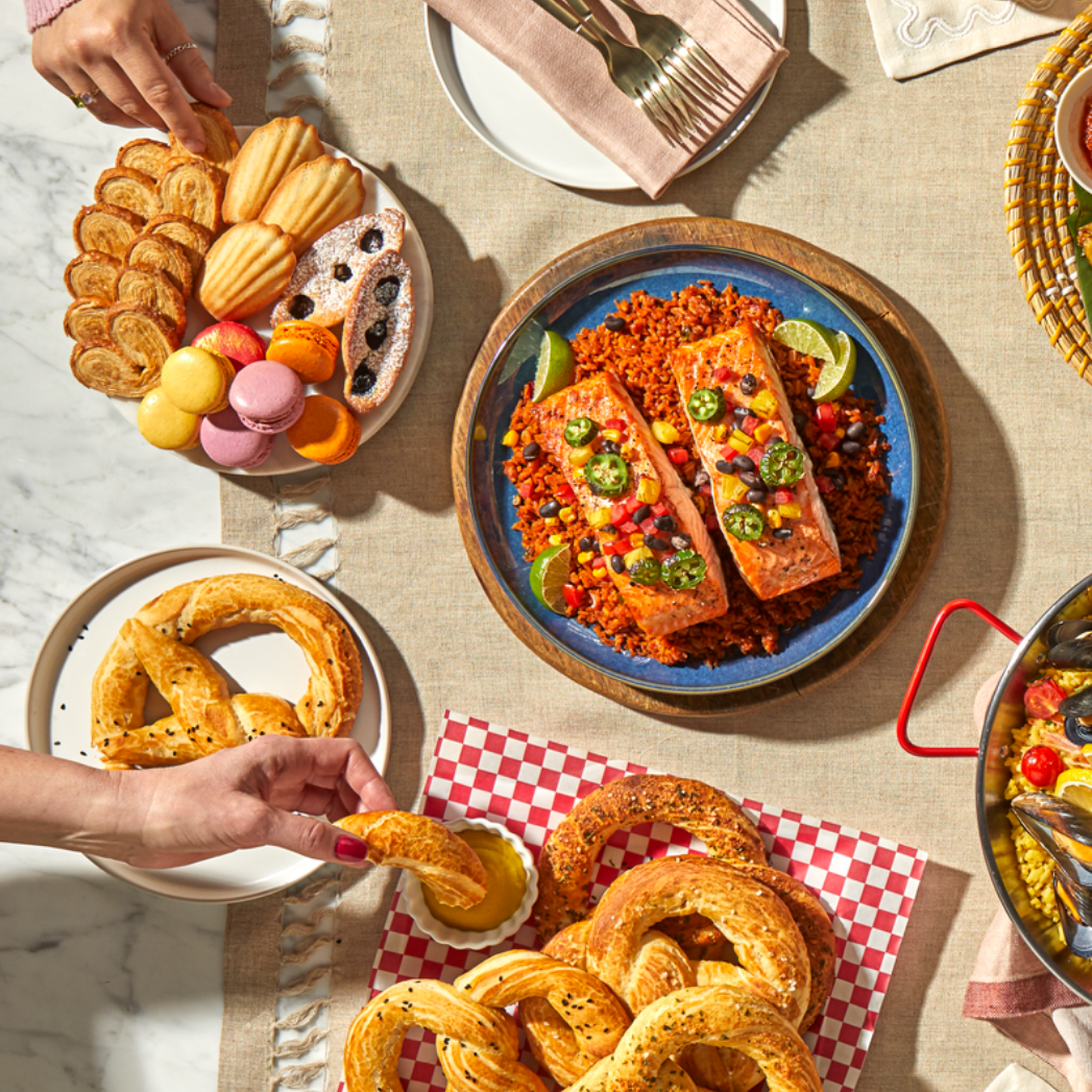 Overhead view of a colorful, festive table spread featuring salmon fillets over seasoned rice with lime wedges, a plate of assorted cookies and macarons, a plate of sesame pretzels with mustard dipping sauce, and a pot of stew partially visible in the corner. A hand reaches for a pretzel, adding a lively, shared-meal atmosphere.