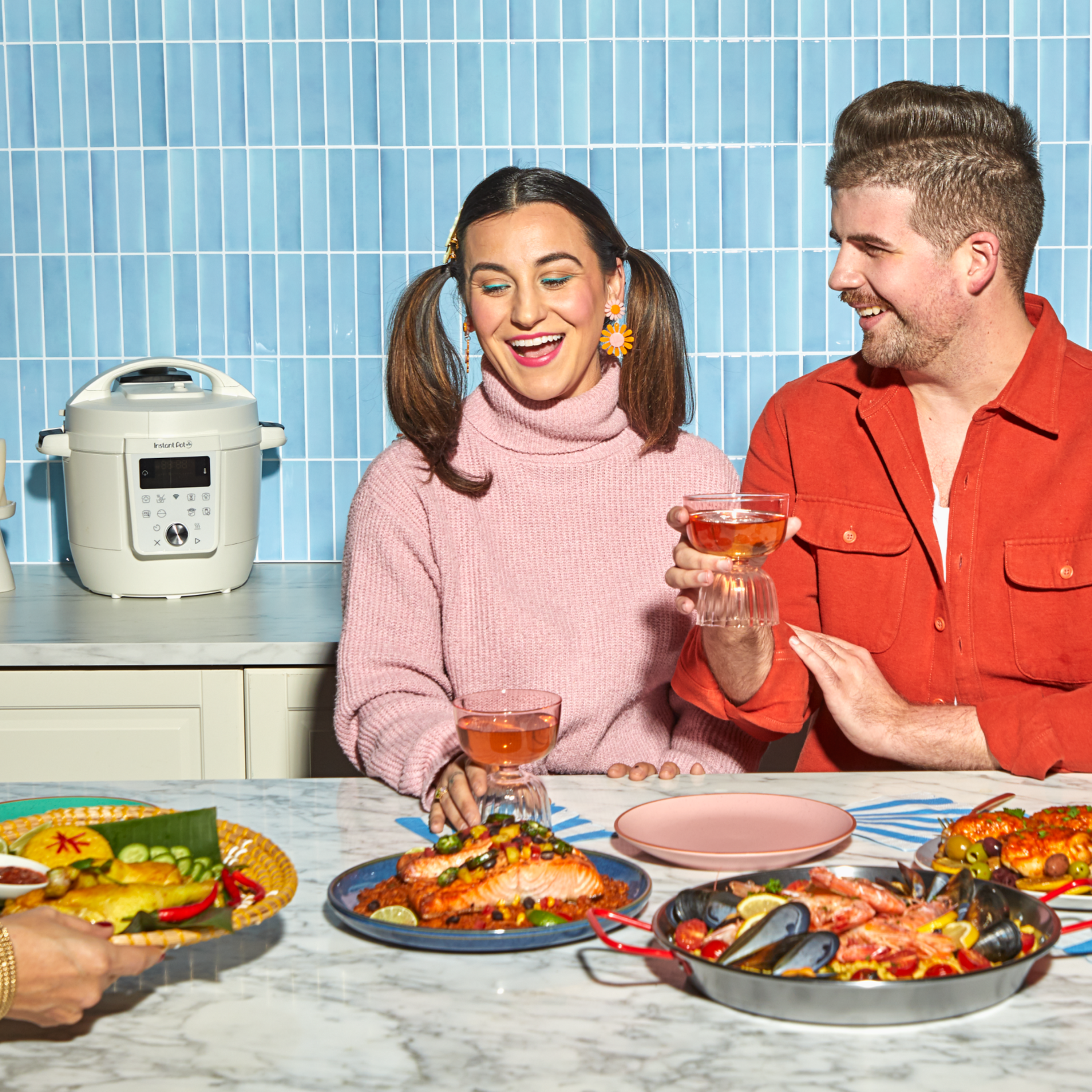 A cheerful group enjoys a colorful seafood meal in a bright kitchen. A woman in a pink turtleneck sweater with pigtails and daisy earrings holds a glass of rosé while smiling at a man in an orange shirt beside her, who also holds a glass. On the table in front of them are plates of vibrant seafood dishes, including salmon, shrimp, mussels, and vegetables. In the background, a white Instant Pot multi-cooker sits on the countertop against a light blue tiled wall.
