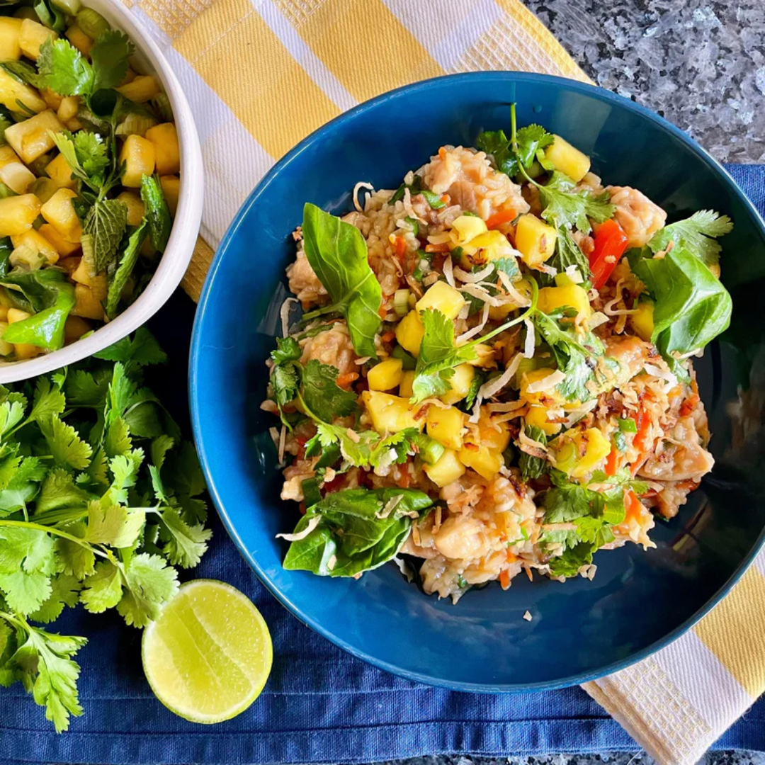 A vibrant bowl of sticky coconut chicken and rice is topped with fresh pineapple salsa, shredded coconut, basil, and cilantro. The dish is served in a deep blue bowl, surrounded by a halved lime, fresh cilantro, and a bowl of extra salsa on a dark granite surface with blue and yellow striped linens.