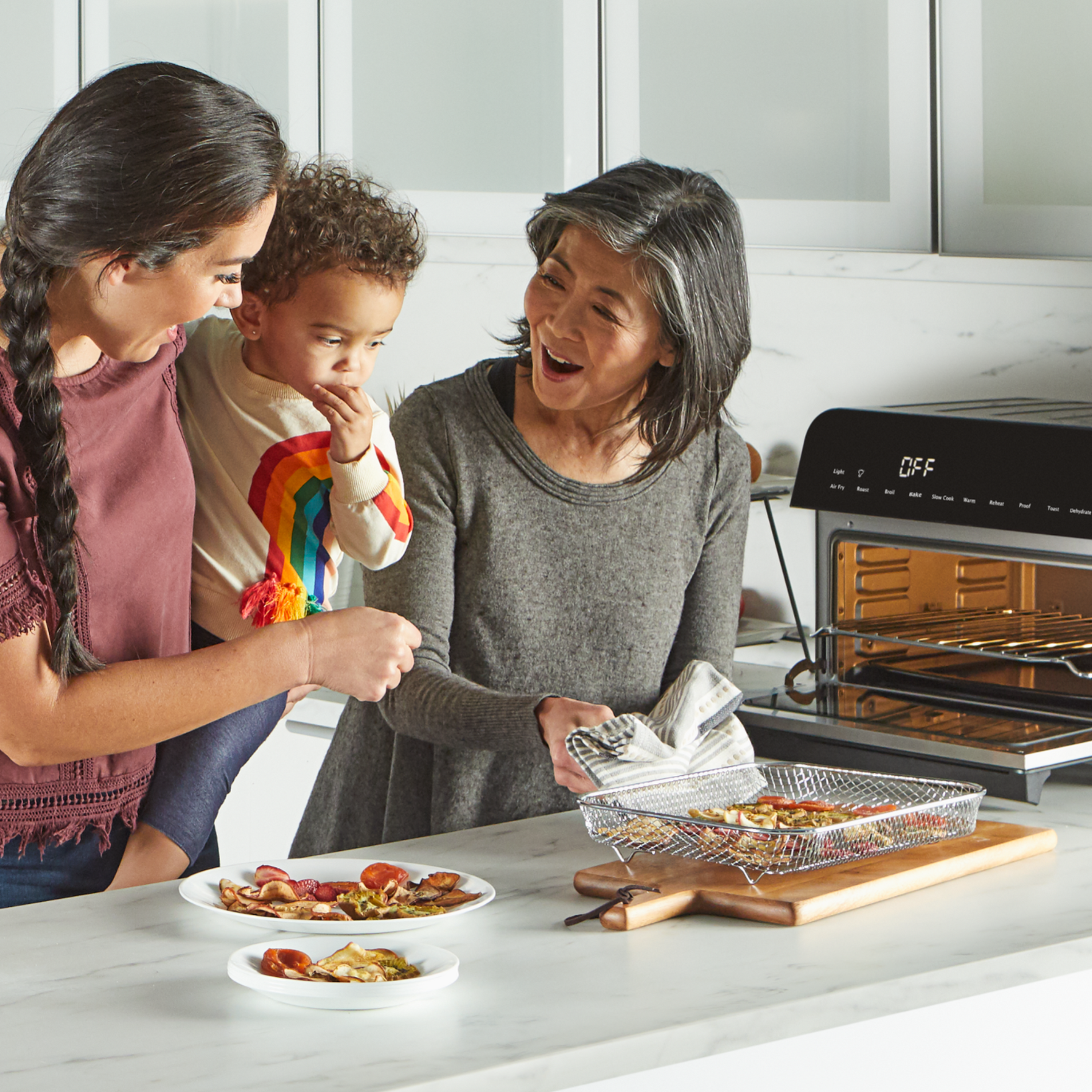 Three generations of a family—an older woman, a young woman, and a toddler—gather in a kitchen, smiling and sharing food from a mesh tray just removed from an Instant Pot® toaster oven. Plates of cooked snacks or mini pizzas sit on the counter in front of them.