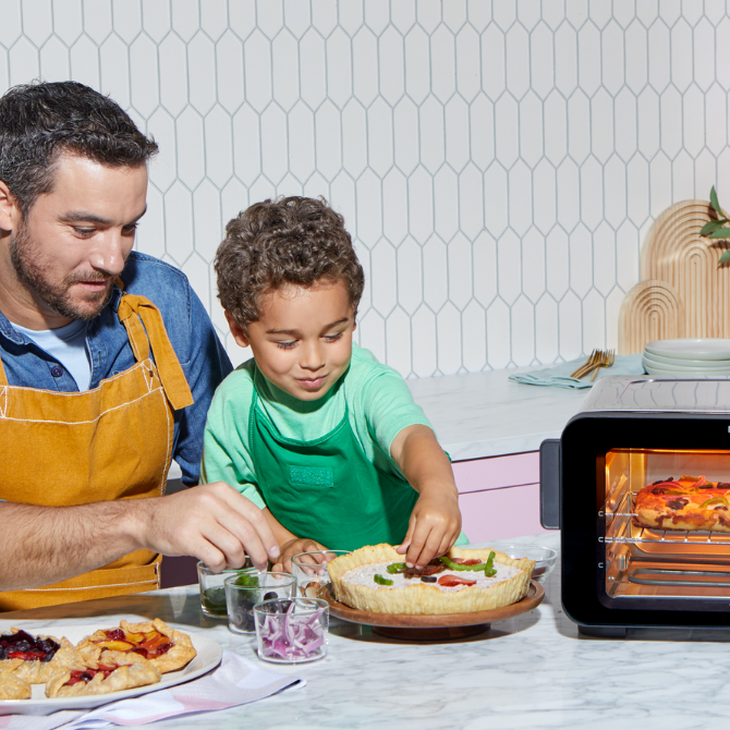 A man and young boy in aprons decorate a tart with fresh toppings at a marble kitchen counter. To the right, the Instant Pot® 6-Slice InstantHeat™ Air Fryer Toaster Oven is baking a pizza inside. The cheerful kitchen scene includes small bowls of ingredients and a plate of finished fruit galettes.