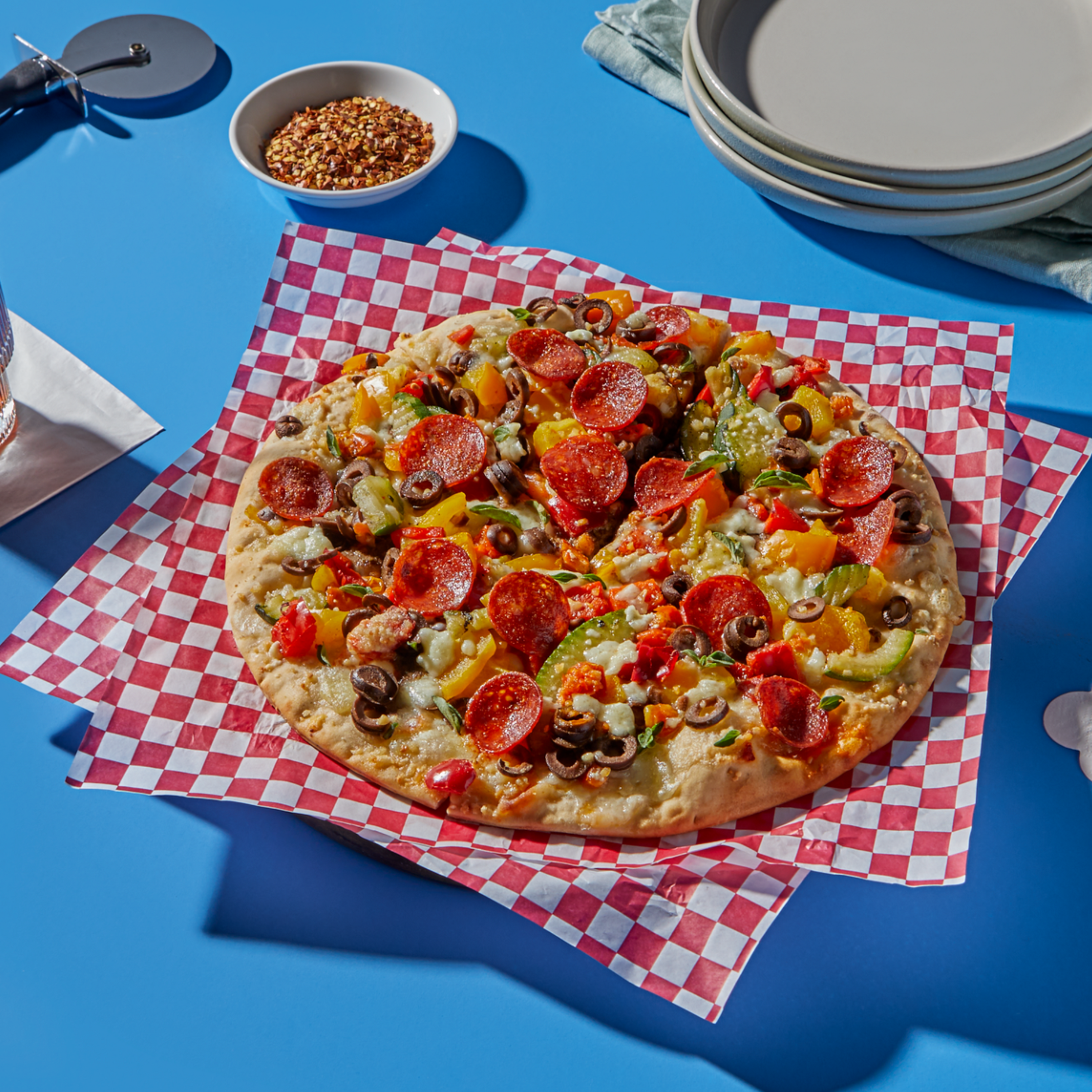 Overhead view of a fully loaded pepperoni pizza with colorful bell peppers, olives, and melted cheese, served on red-and-white checkered paper over a bright blue surface. A stack of plates, pizza cutter, and bowl of red pepper flakes are nearby.