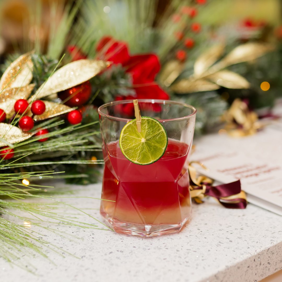 A festive red holiday drink garnished with a dehydrated lime slice sits in a faceted glass on a countertop, with evergreen décor, red berries, and gold ribbon blurred in the background.