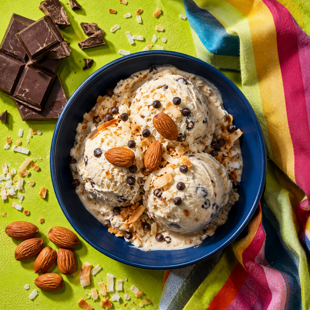 Three scoops of coconut milk ice cream with chocolate chips, toasted coconut, and almonds in a navy blue bowl on a lime green surface with chocolate pieces and a striped napkin.