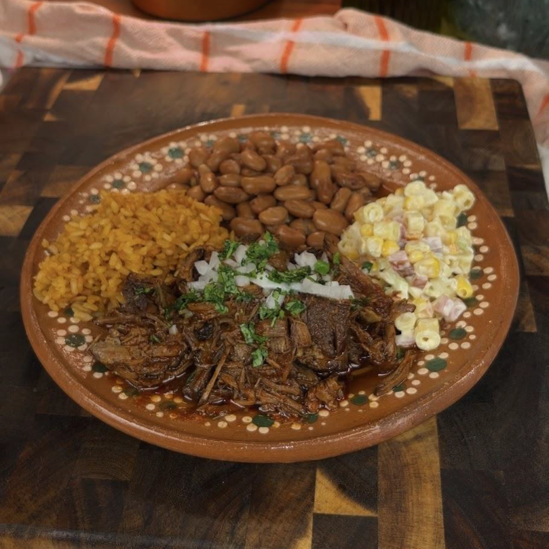 A rustic brown plate with shredded barbacoa topped with chopped onions and cilantro, served alongside Mexican rice, pinto beans, and a creamy corn salad. 